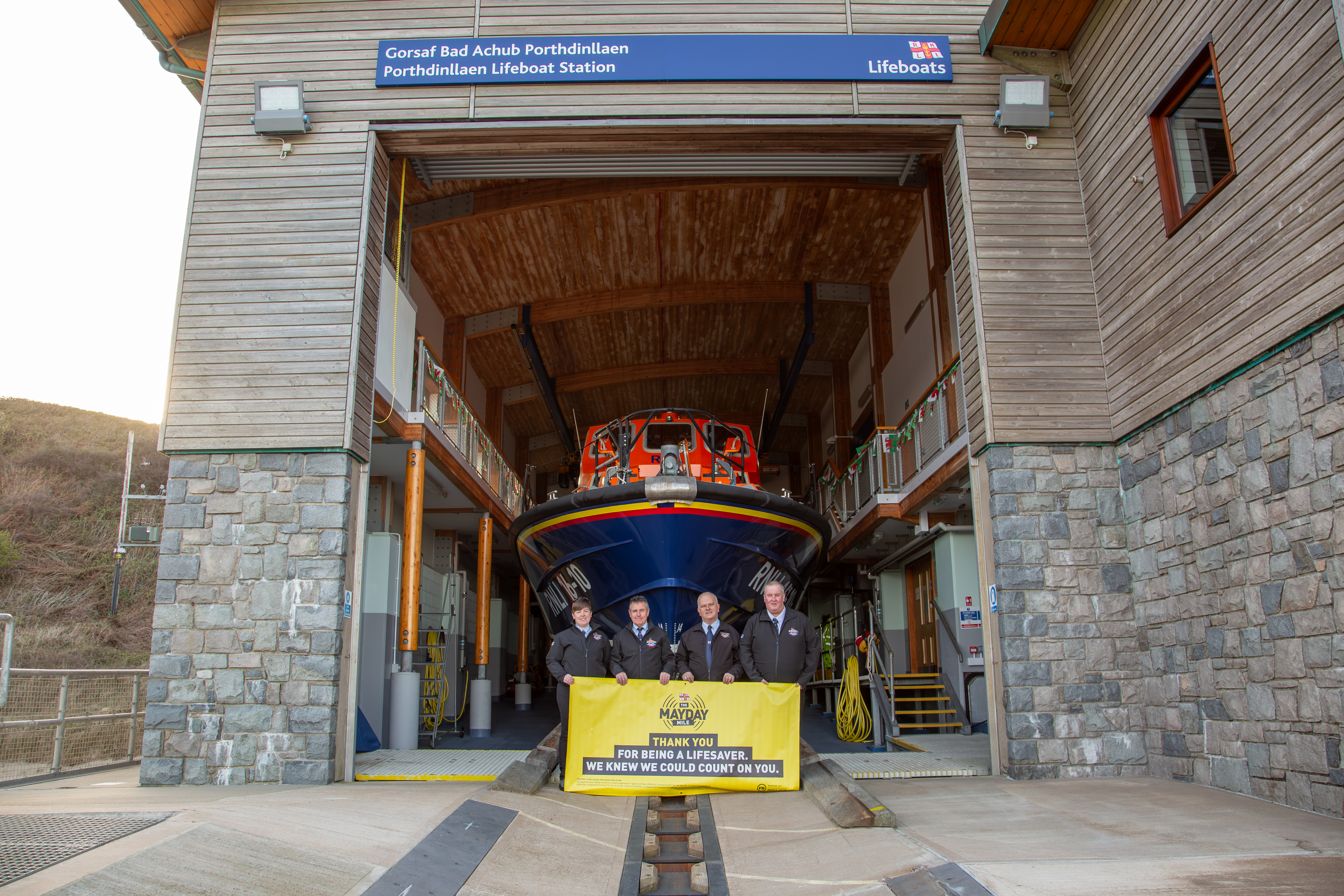 Landscape image of the four volunteers holding Mayday banner with a backdrop of Porthdinllaen lifeboat station.
