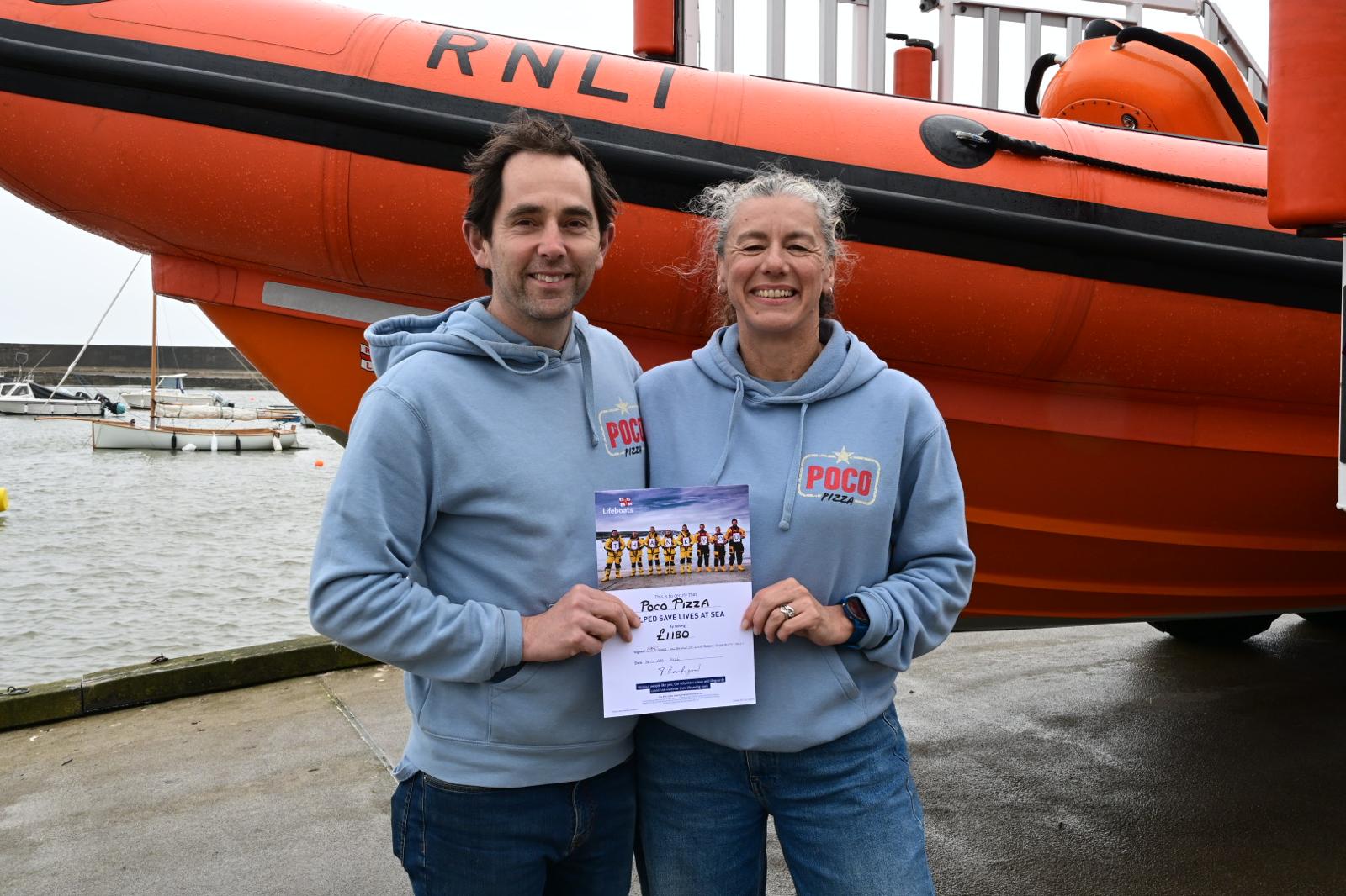 Two people in blue tops with certificate in front of lifeboat
