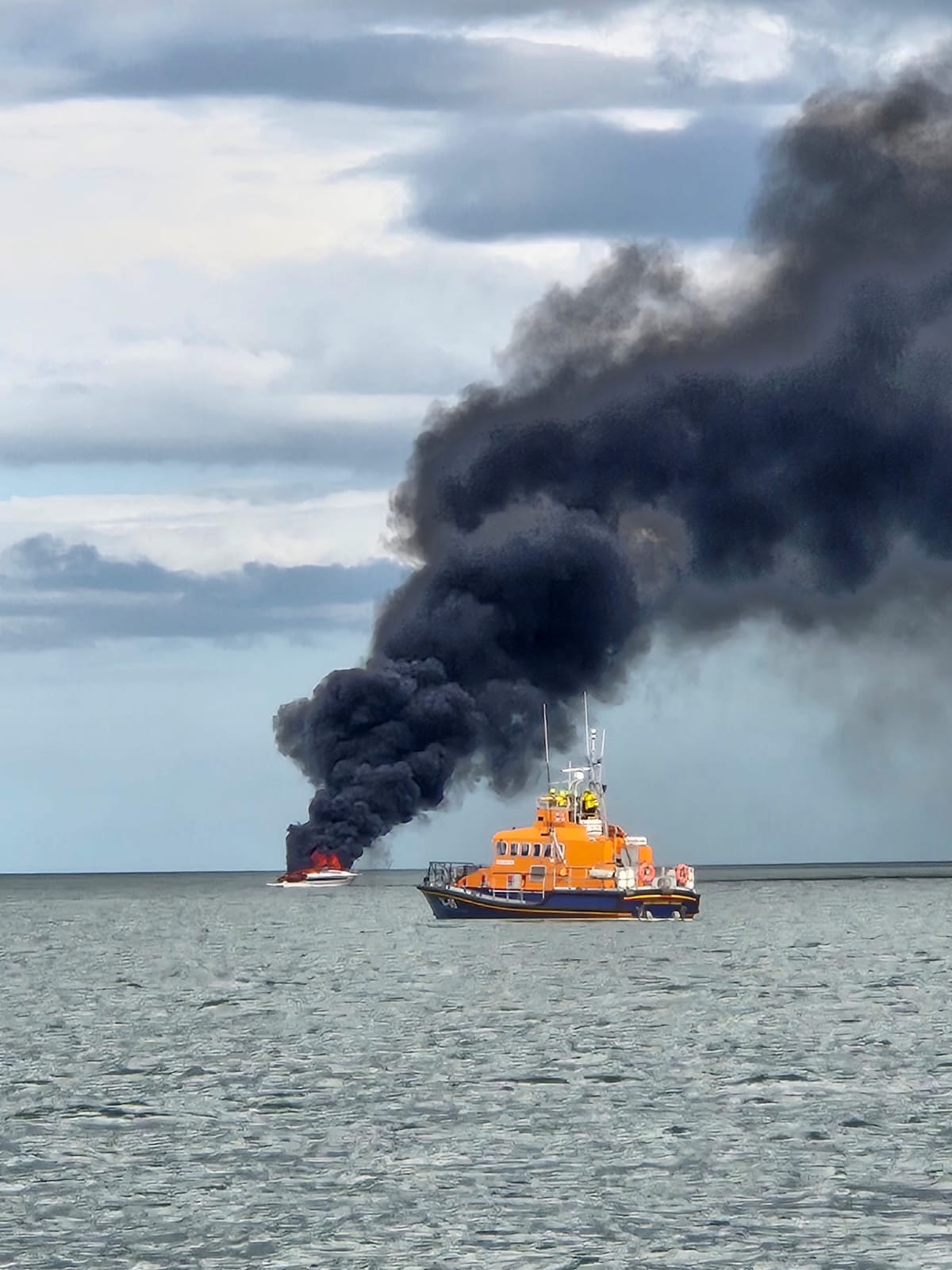 Lifeboat in calm water with a vessel on fire in the background. Huge black smoke plumes in sky. Cloudy skies