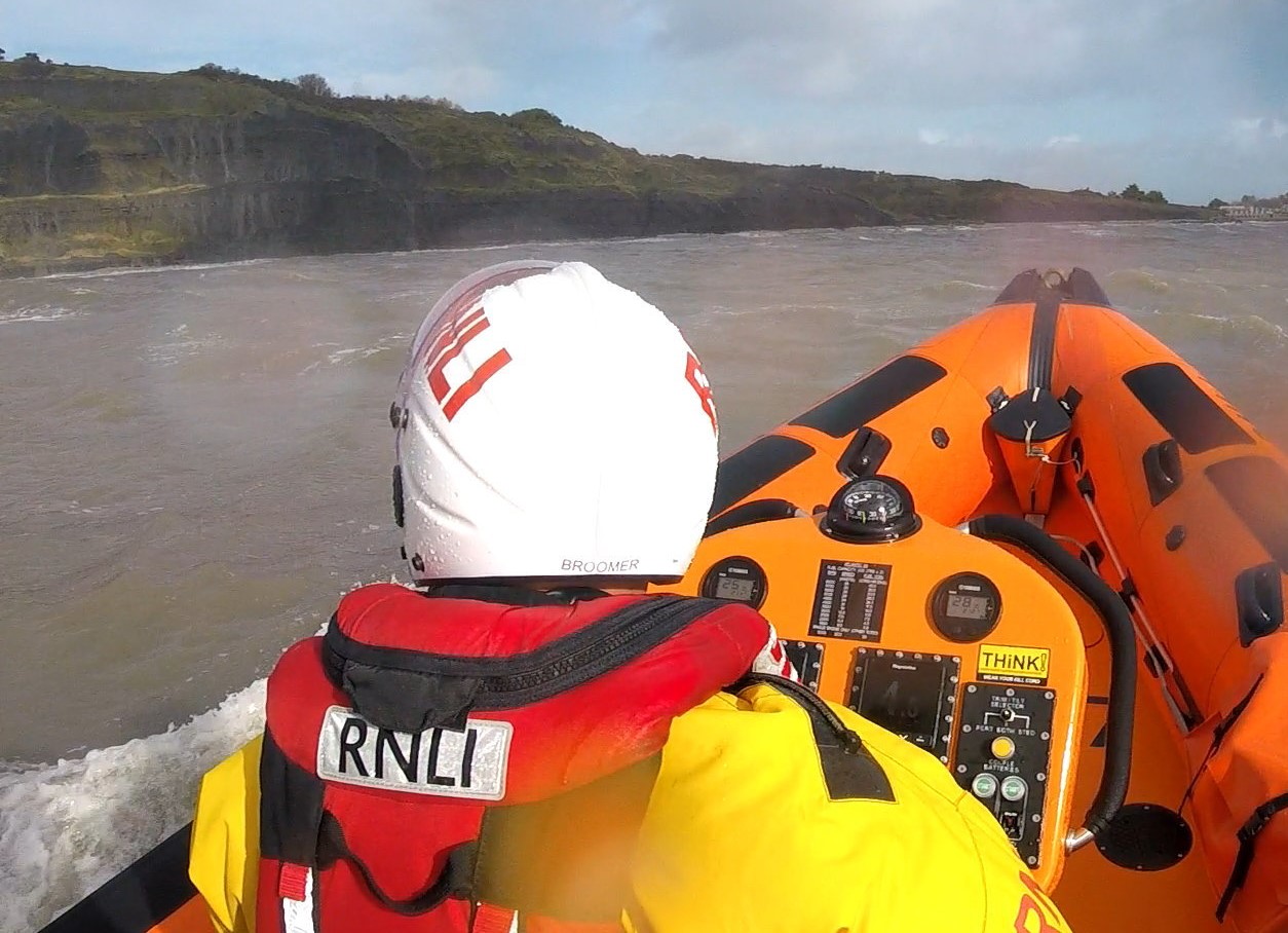man in yellow and red lifeboat clothing on orange boat with coast in the background