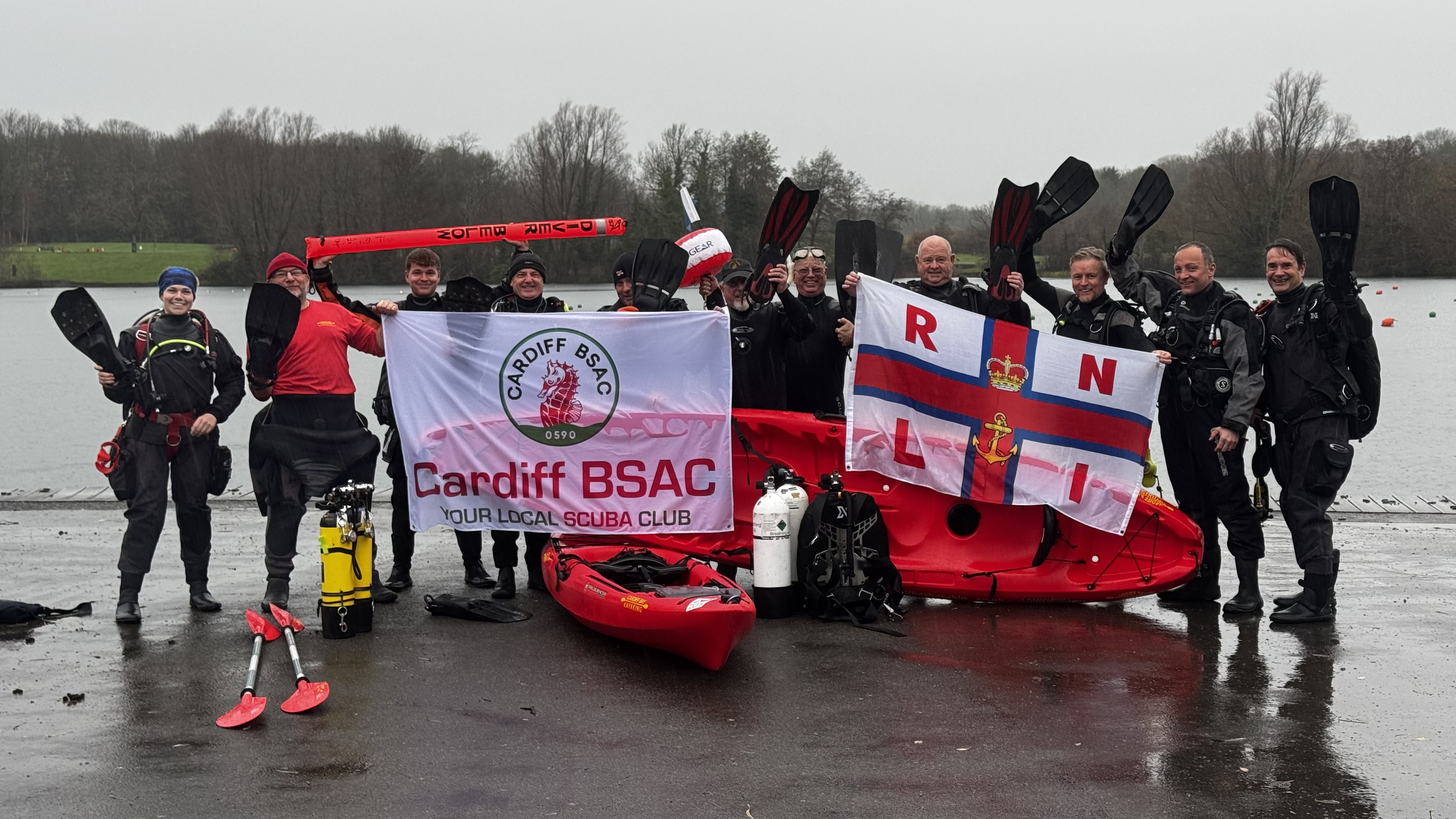 Group image of the Cardiff BSAC team with RNLI and their club flag. Stood next to kayaks. Backdrop of trees and lake.
