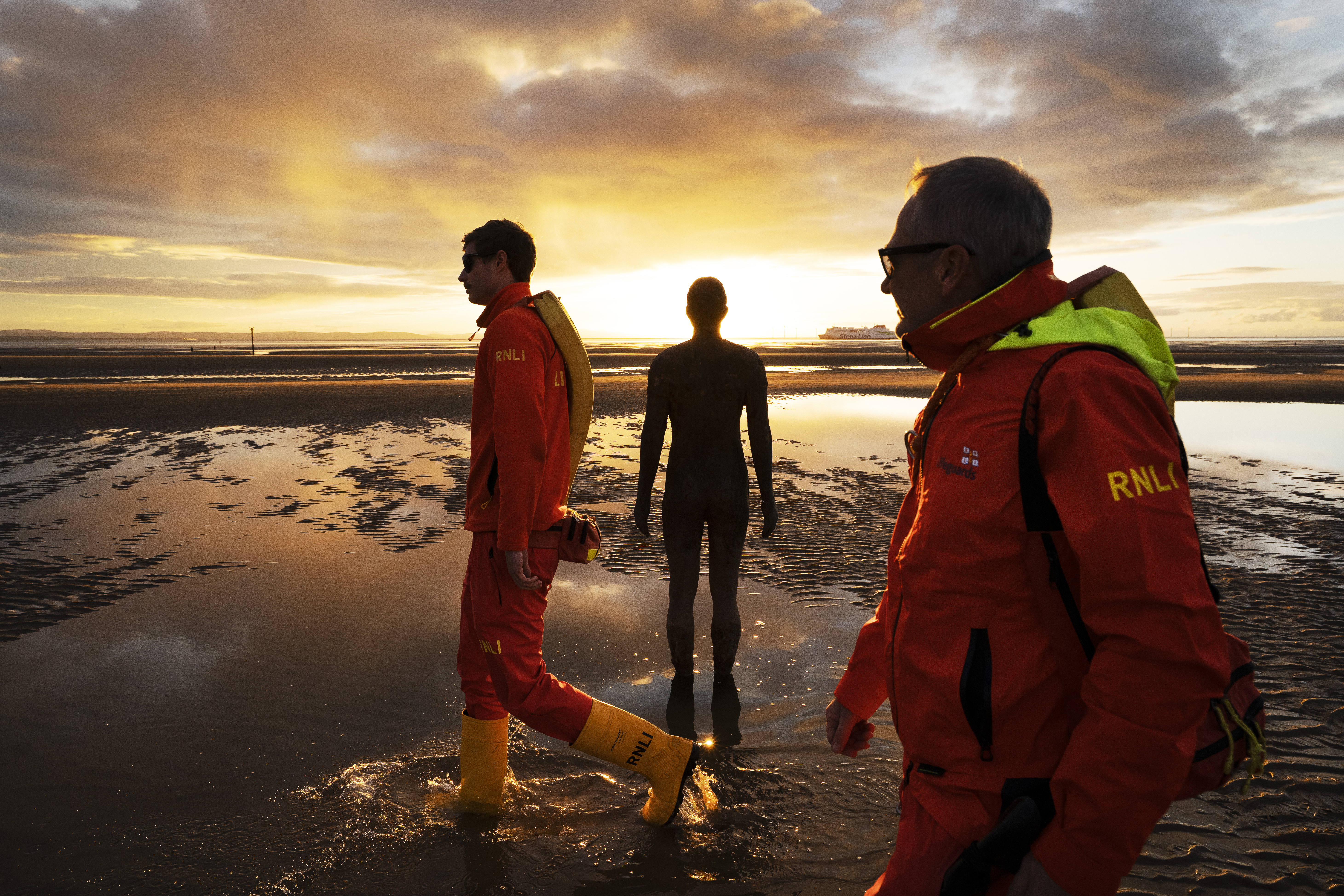 Landscape sunset image of Crosby beach with beach lifeguards walking past one of the metal statues.