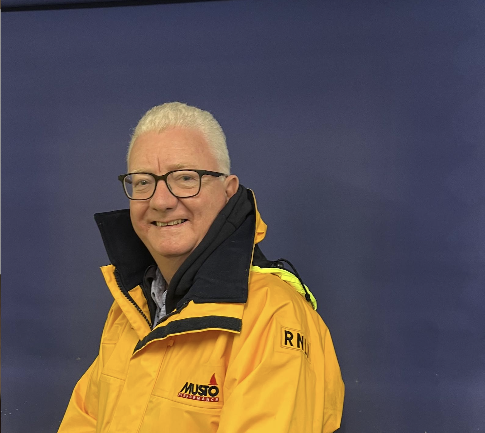 Greg, looks over his left shoulder wearing a yellow RNLI jacket, smiles at the camera