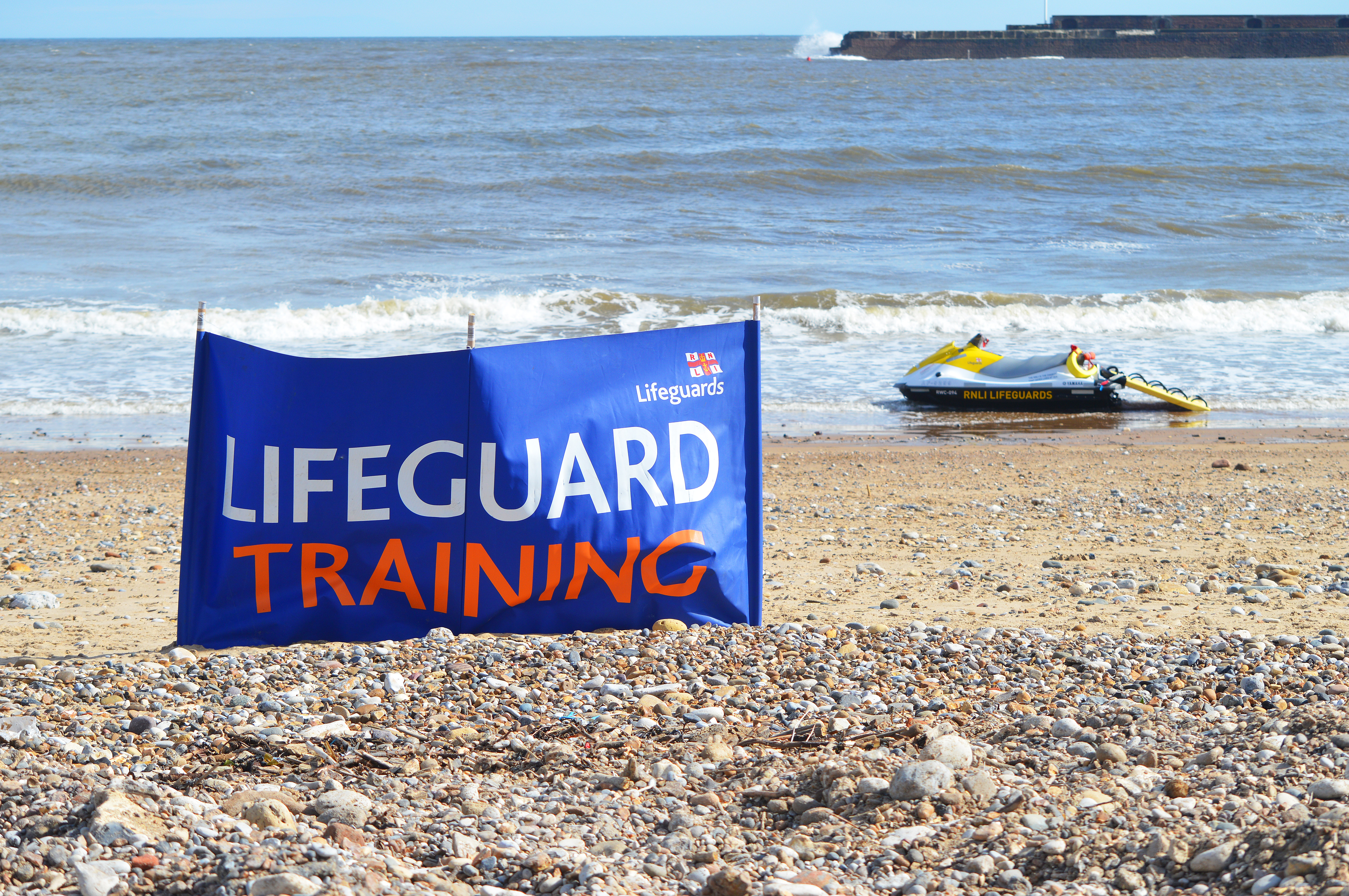 A banner with the wording 'Lifeguard Training' upon it has been fixed into place on a beach. In the background - where the shore meets the sea - a lifeguard watercraft is on standby.