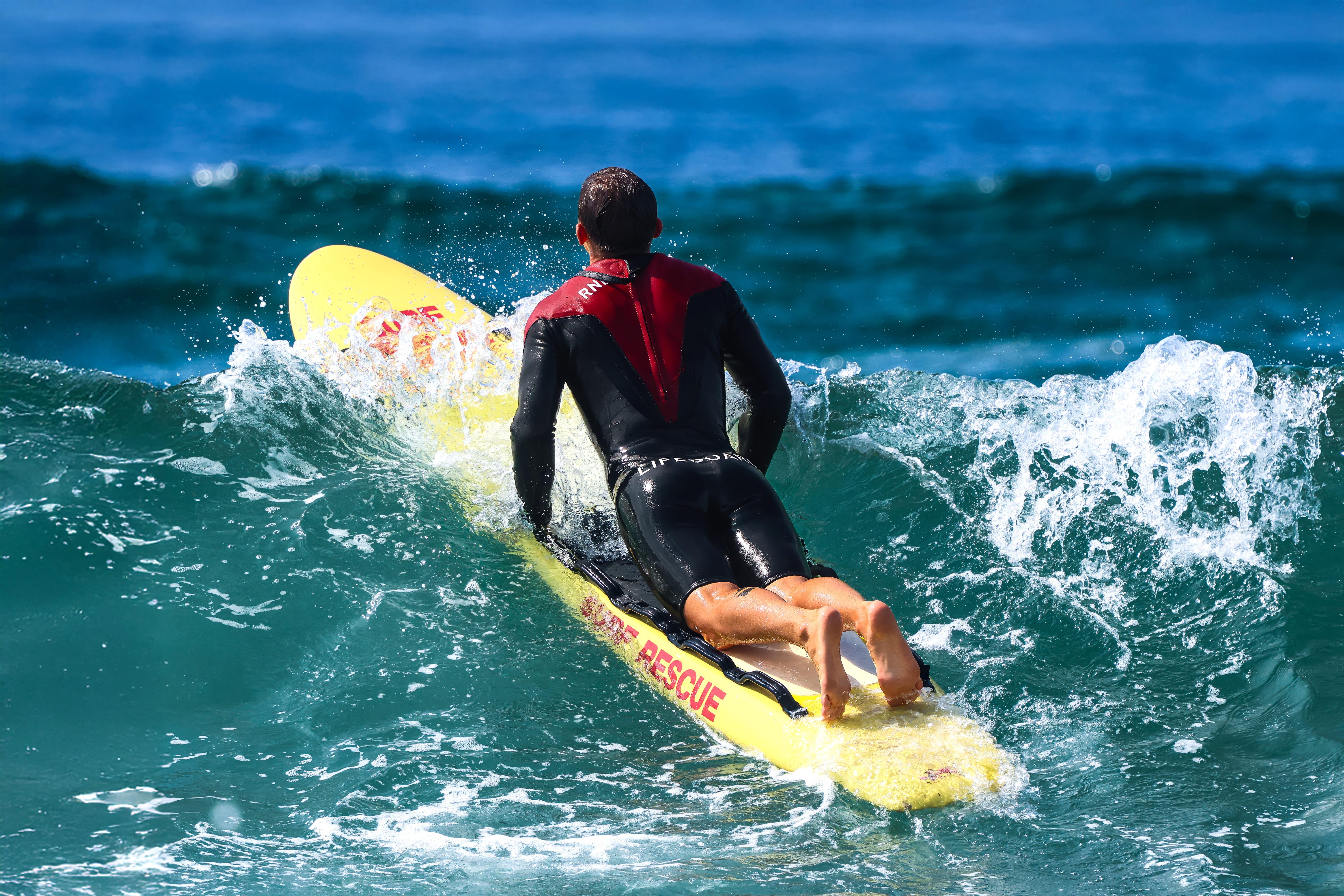 A RNLI lifeguard rides the surf upon a yellow rescue board. The lifeguard is viewed from behind and he is propped up - from the waist upwards - against the rescue board as he looks out to sea.