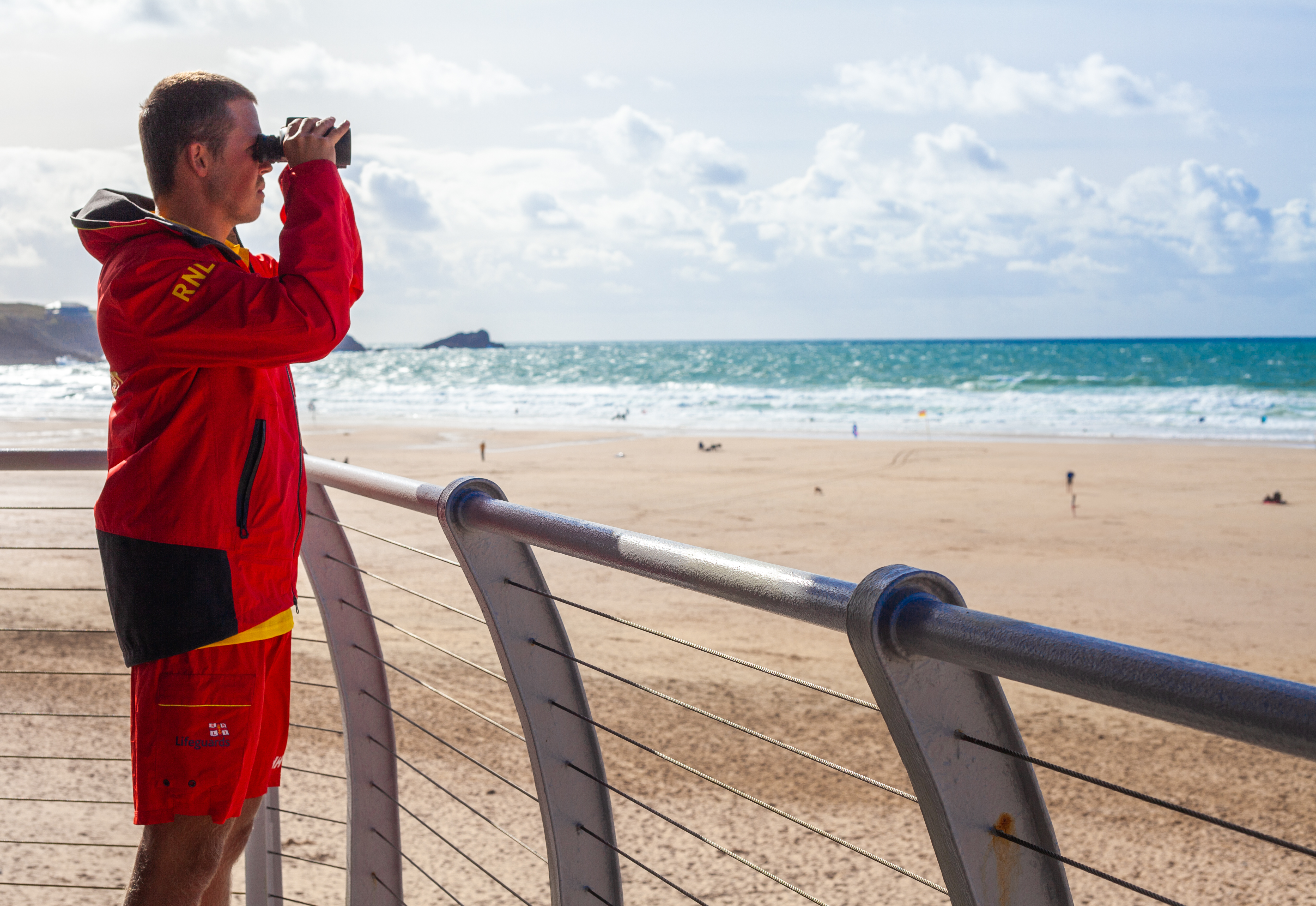 A RNLI lifeguard observes beach goers through binoculars from behind a railing overlooking the coast. The weather is dry and sunny.