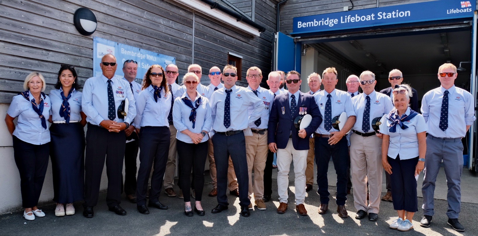 Crew from Bembridge RNLI lined up outside Bembridge Lifeboat station together, facing the camera in their blue shirt uniform
