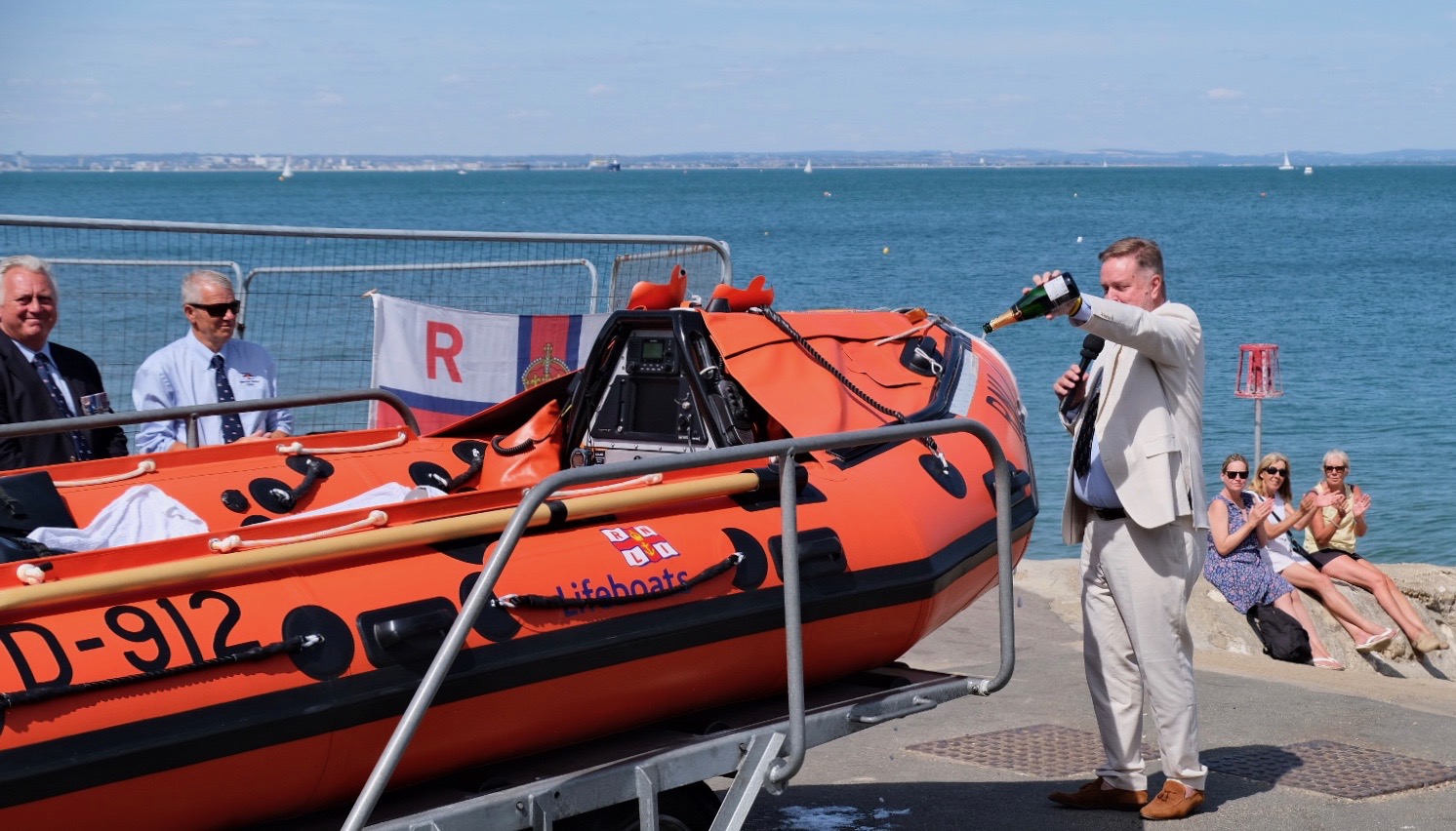 High Sherriff pouring champagne on the nose of the inshore lifeboat for the official christening, beach in the background with a few spectators