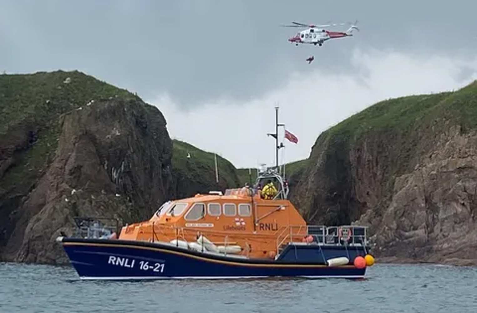 Fraserburgh RNLI ALB 16-21 John Buchanan Barr in foreground as casualty is winched aboard Coastguard Rescue Helicopter R851 from inaccessible gully, high green cliffs in the background.