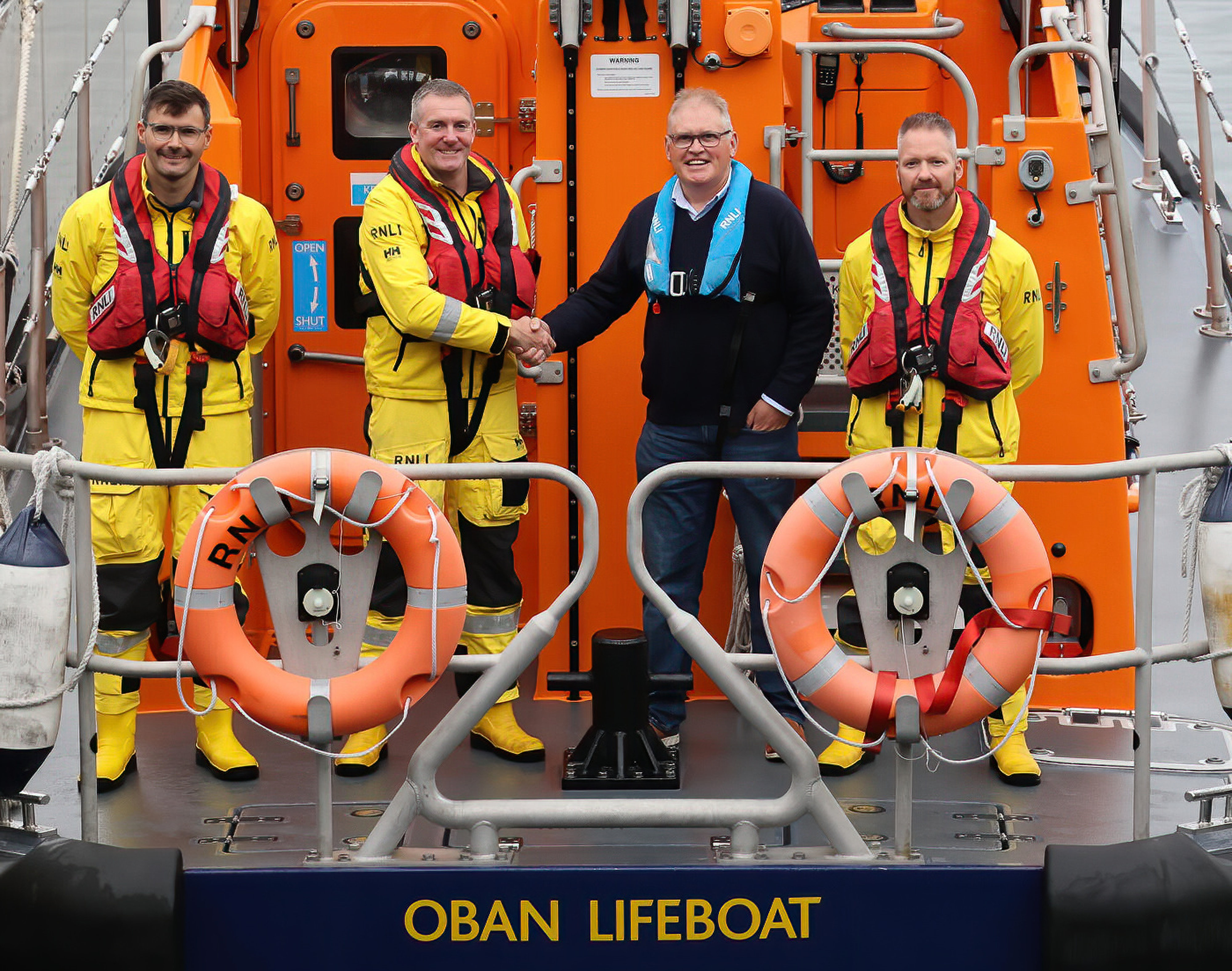 Three lifeboat crew members in their yellows are joined by Brian Clarke on the aft deck of the Shannon class lifeboat. Brian and Ally are shaking hands.