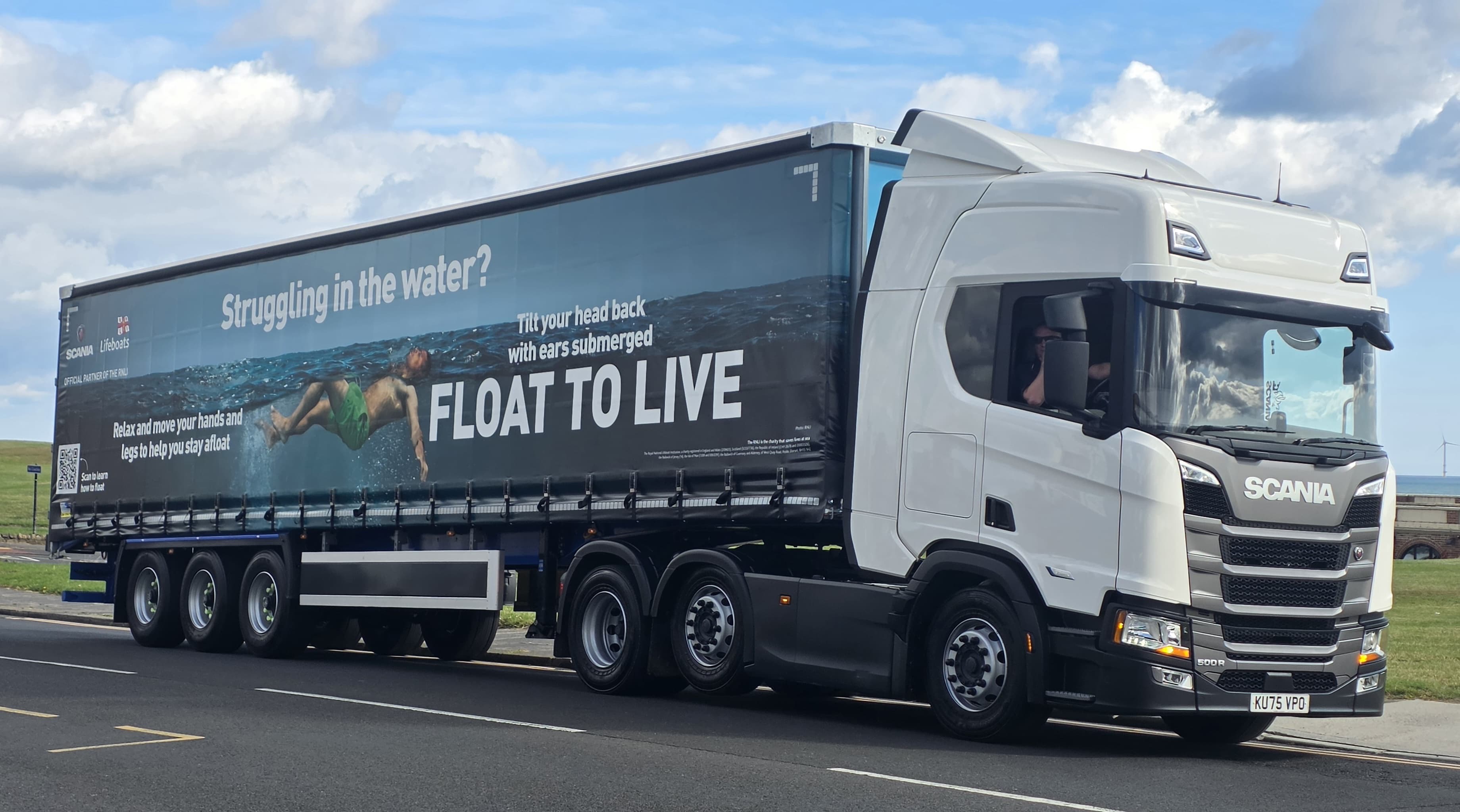 Image shows a Scania truck and trailer parked on a road. The cabin is to the right of the image, the rear of the trailer is to the left. The trailer has a RNLI Float to Live campaign image on it, running the full length of the trailer. In the background, some grass and the sea can be seen. The weather is bright and dry with some clouds in the sky.