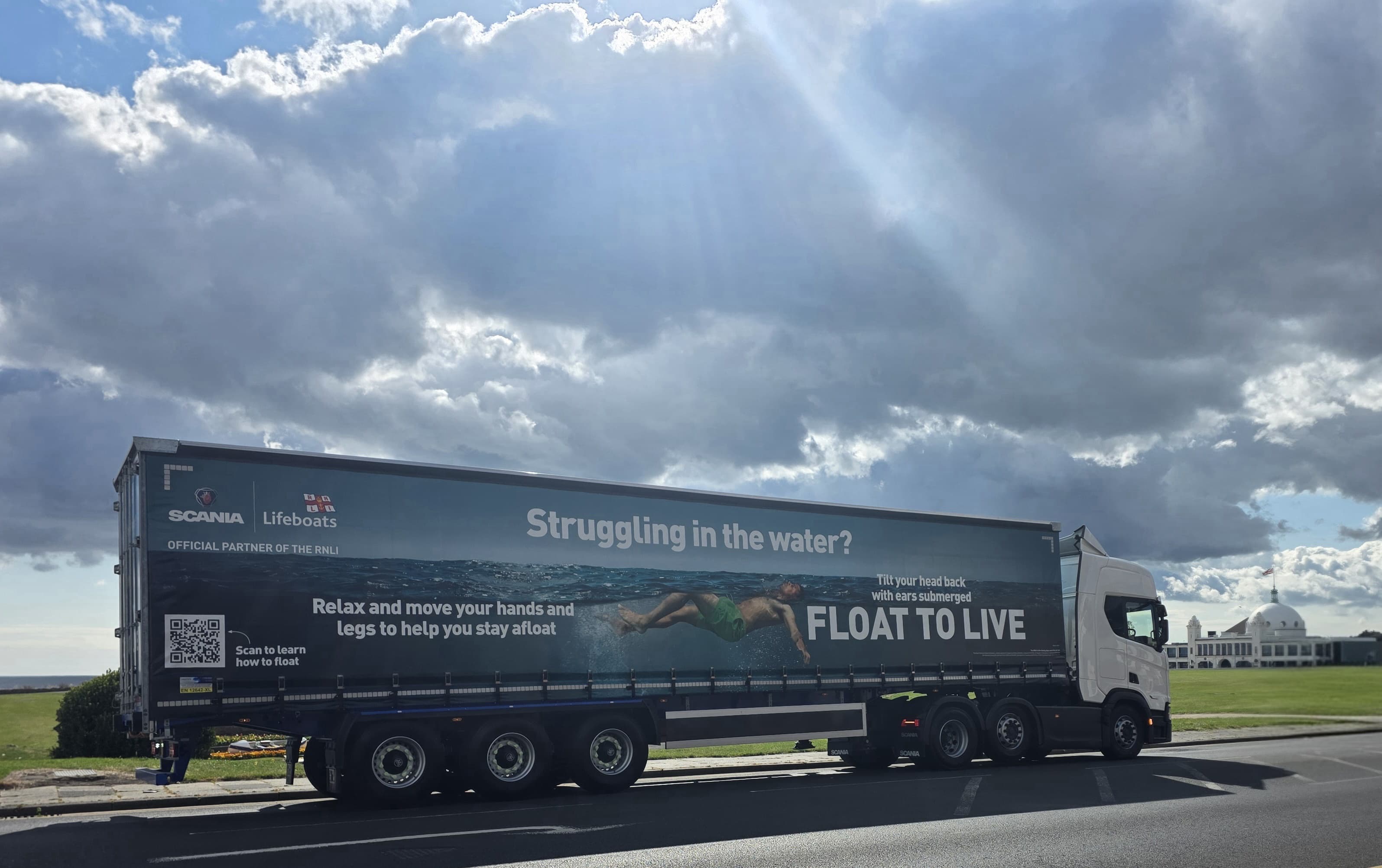 Image shows a Scania truck and trailer parked on a road. The cabin is to the right of the image, the rear of the trailer is to the left. The trailer has a RNLI Float to Live campaign image on it, running the full length of the trailer. In the background, some grass, a building known as Spanish City and the sea can be seen. The weather is bright and dry with some clouds in the sky.