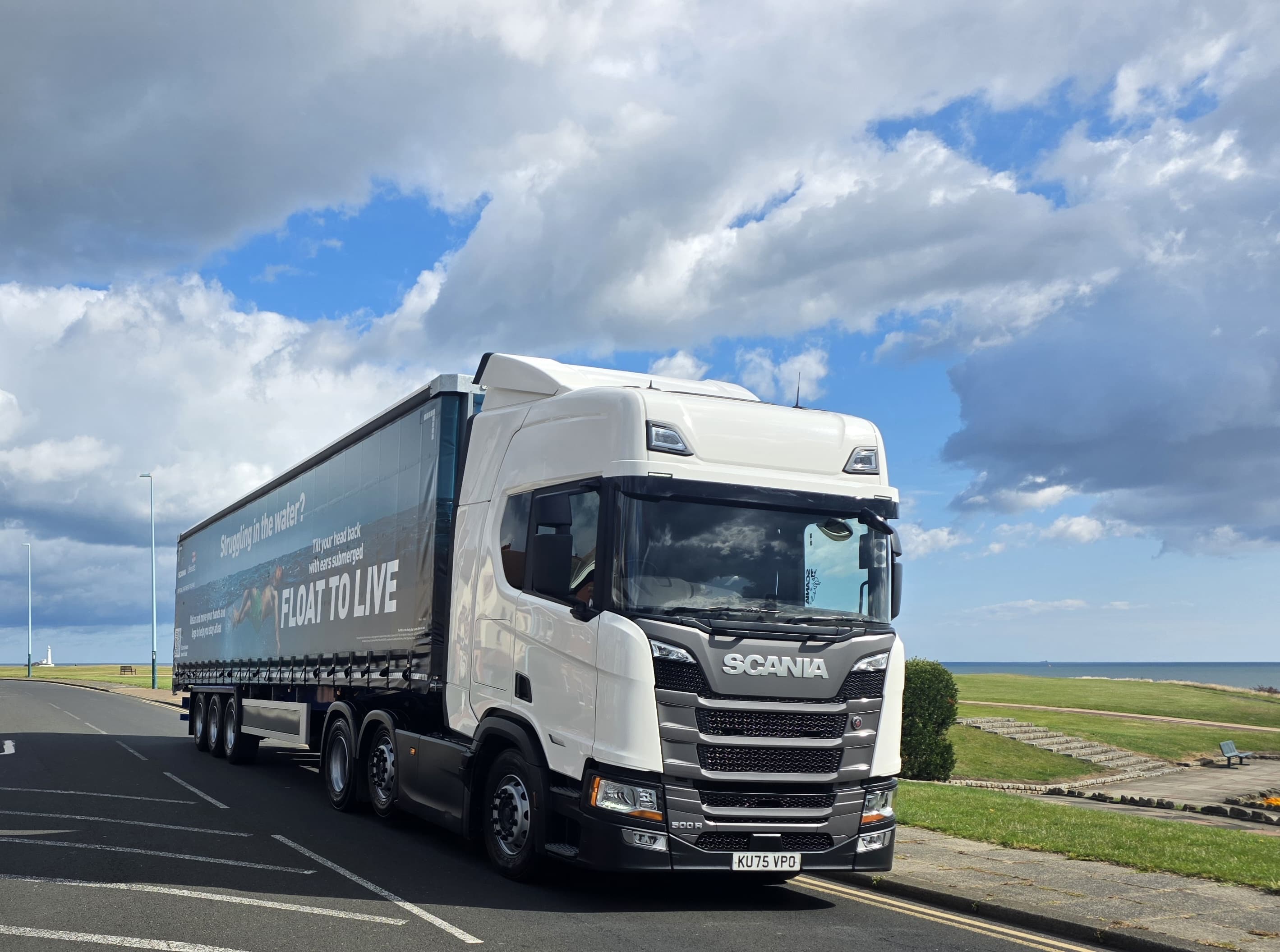 Image shows a Scania truck and trailer parked on a road. The cabin is pointing towards the camera, the rear of the trailer is further behind. The trailer has a RNLI Float to Live campaign image on it, running the full length of the trailer. In the background, some grass, paths and the sea can be seen. The weather is bright and dry with some clouds in the sky.
