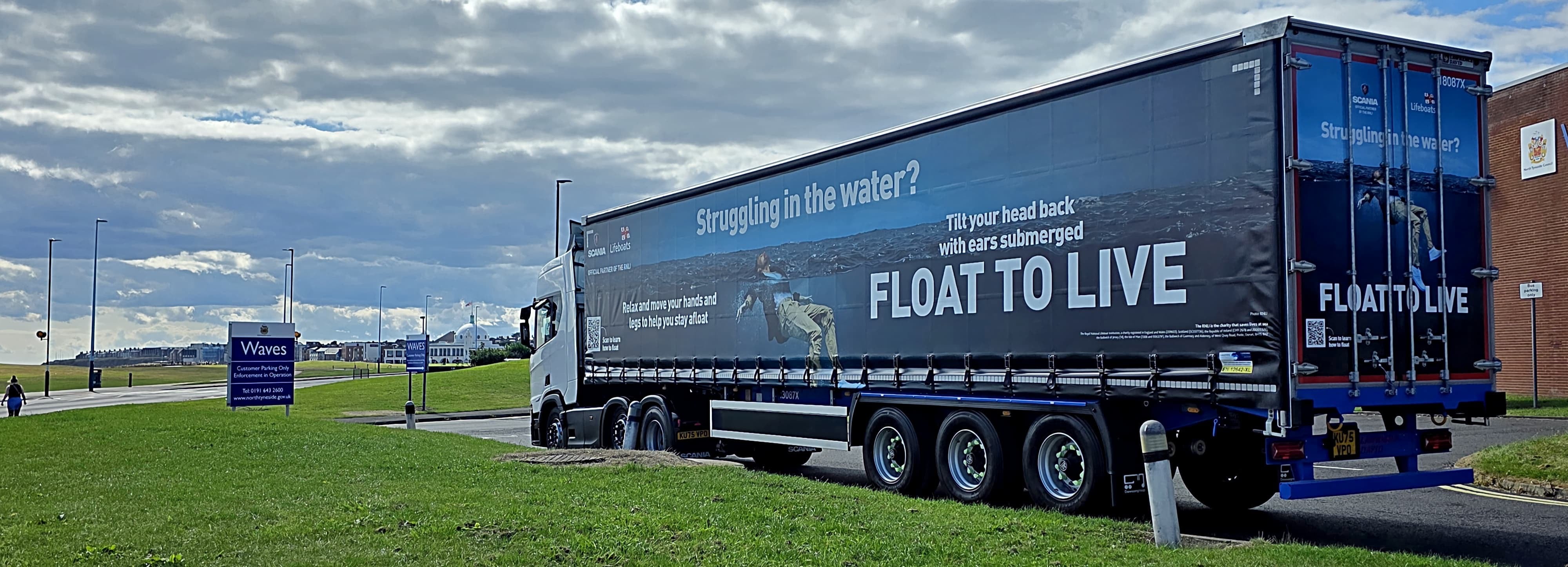 Image shows a Scania truck and trailer parked at the edge of a car park for the Waves leisure centre. The rear of the truck is pointing towards the camera, the cabin of the truck is facing away. The trailer has a RNLI Float to Live campaign image on it, running the full length of the trailer. In the foreground, some grass and a sign for the leisure centre can be seen. In the background, some of the leisure centre can be seen. The weather is bright and dry with some clouds in the sky.