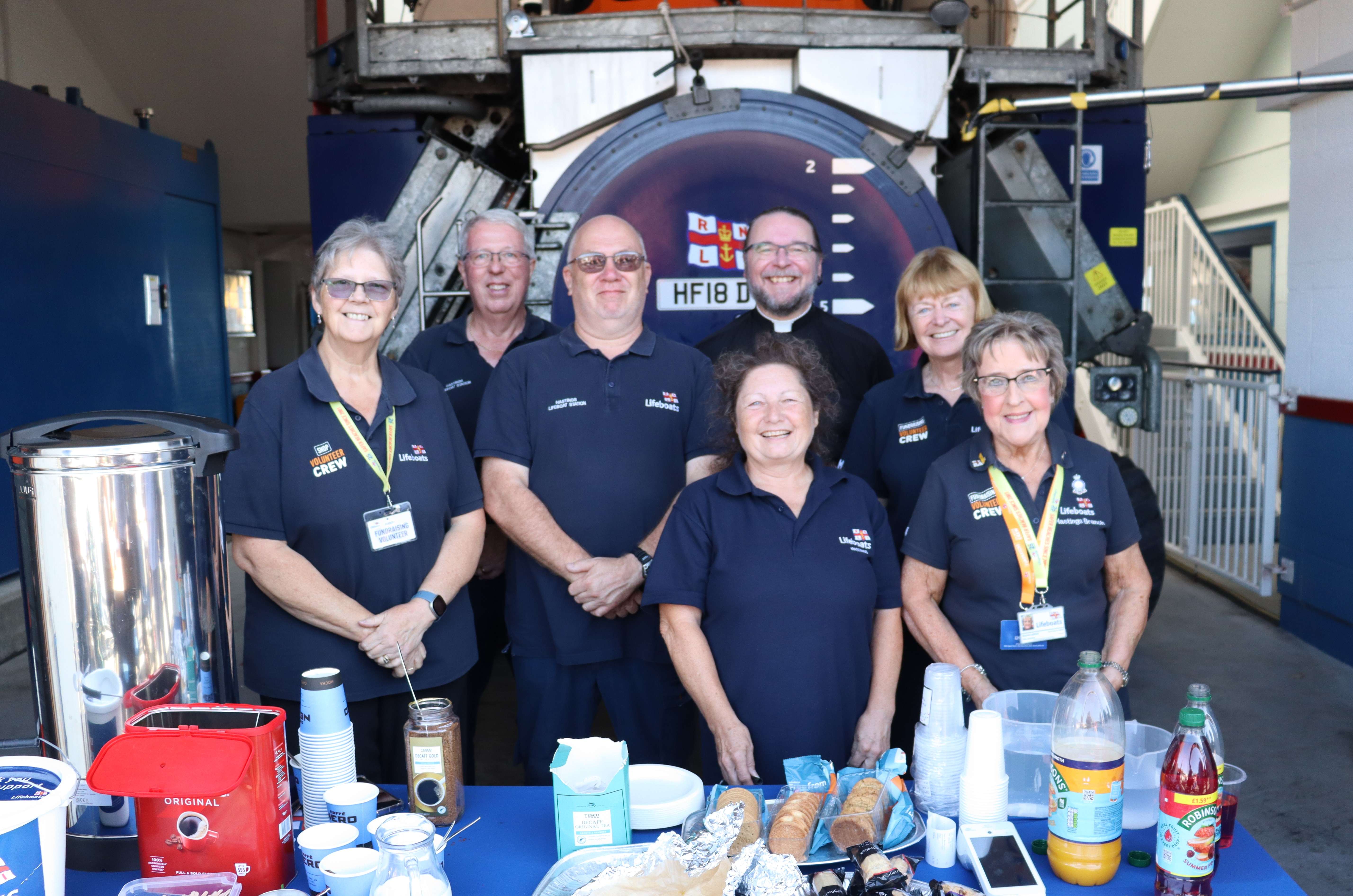 Rev Mat Phipps is surounded by RNLI volunteers, with refreshments on a table in the foreground and the lifeboat SLRS (launch tractor) as a backdrop