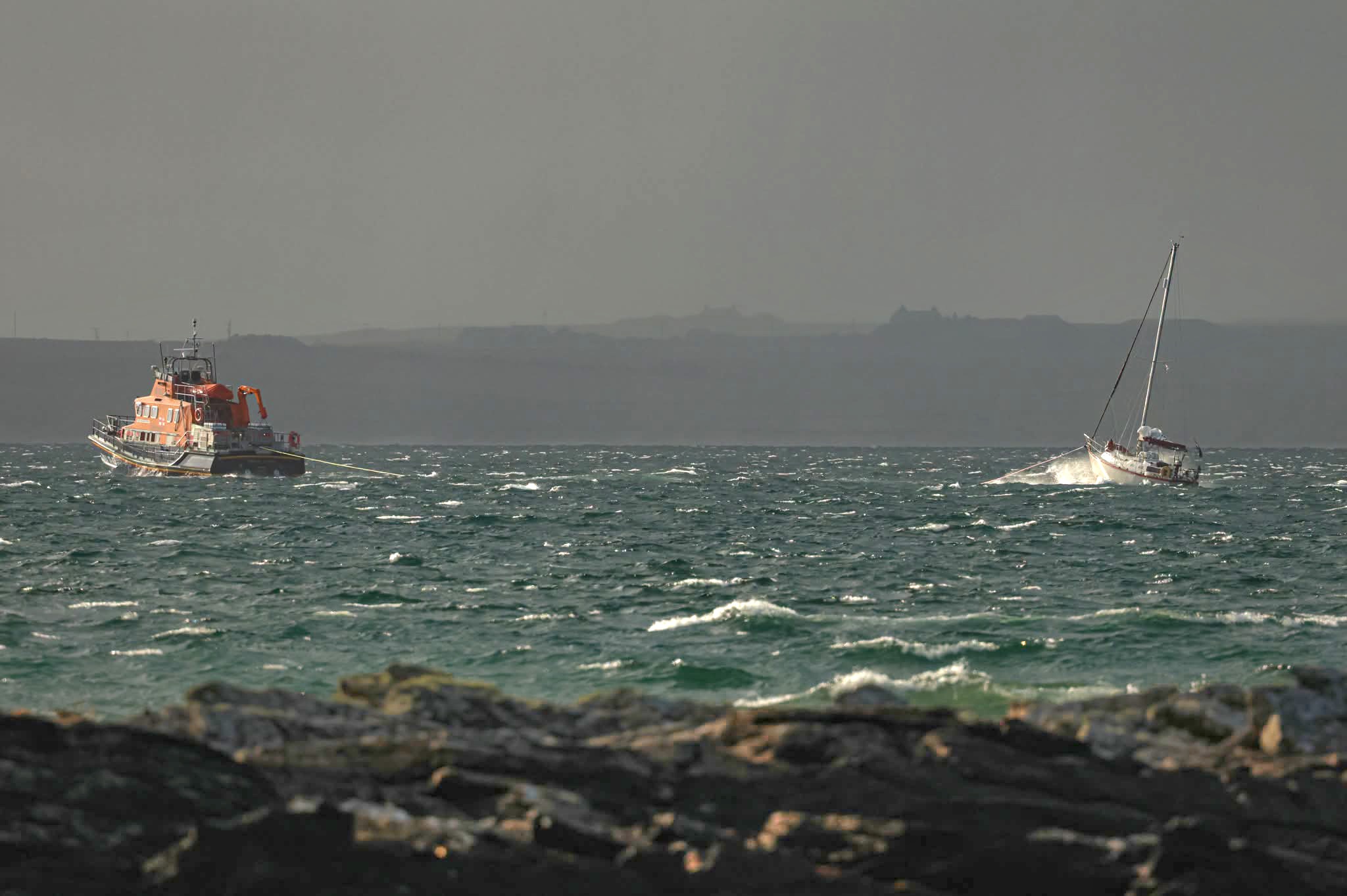 Lifeboat towing small yacht through rough seas, the shoreline is in the foreground and land in the background.