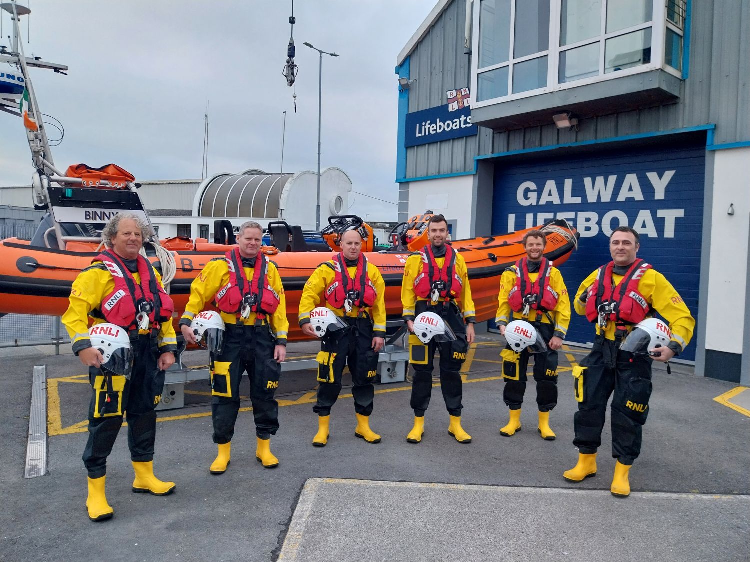 Six crew in drysuits and lifejackets in a row holding helmets and standing in front of the inshore lifeboat in the yeard in front of the lifeboat station.