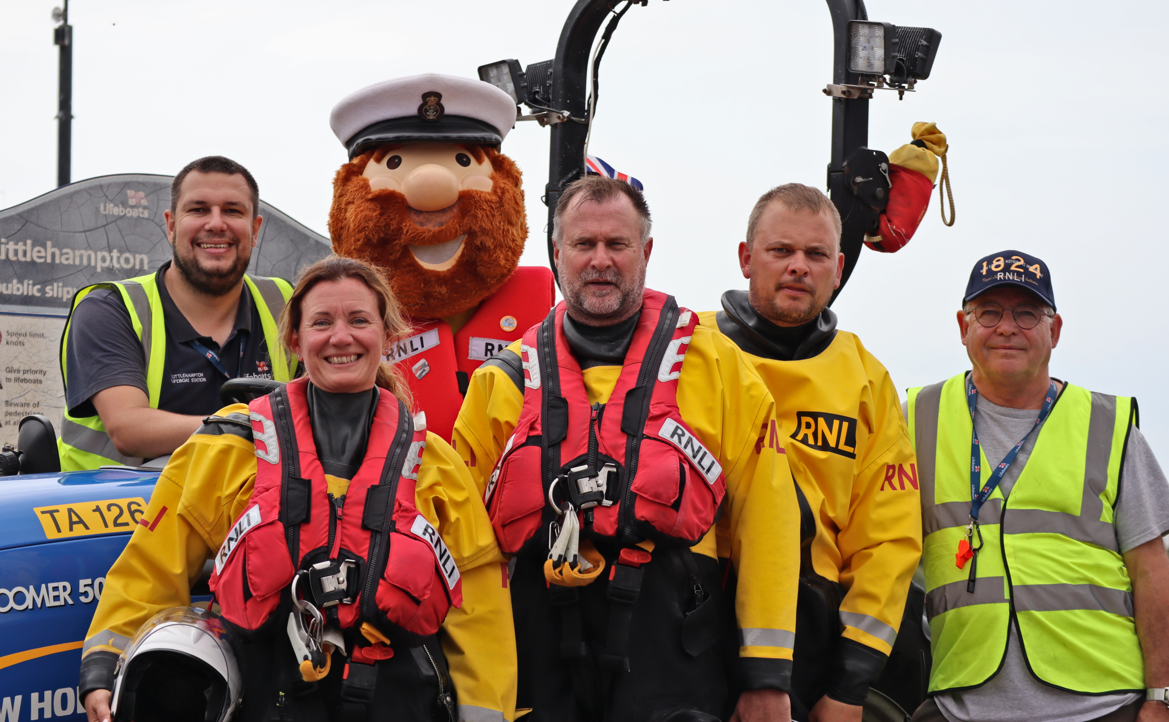 Five volunteers plus an RNLI mascot pose for a group photo by a tractor