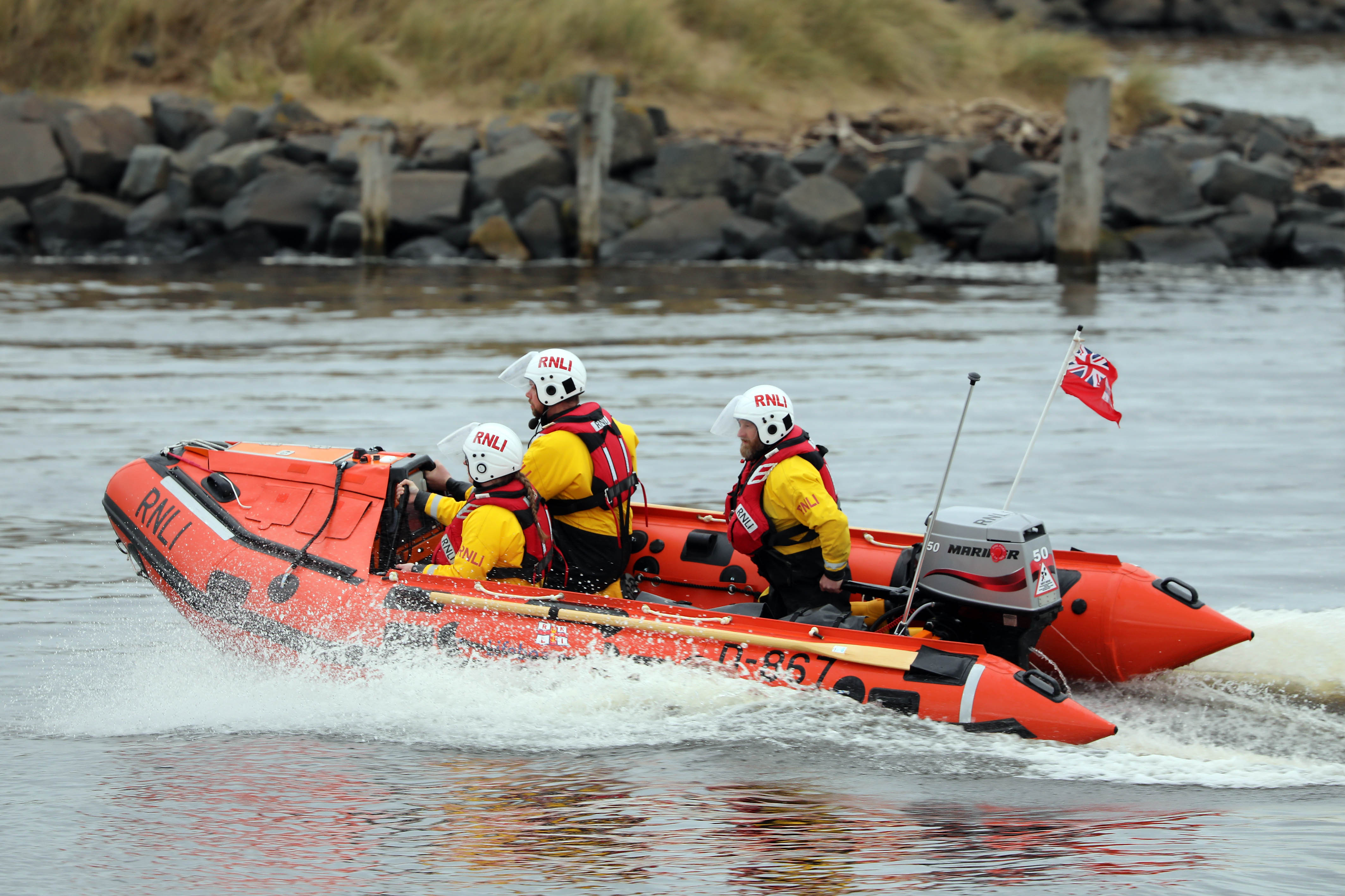 This image shows a RNLI in-shore lifeboat making progress in the water within an estuary. There are three crew onboard the lifeboat. Some splashes of water can be seen coming off the sides of the lifeboat.