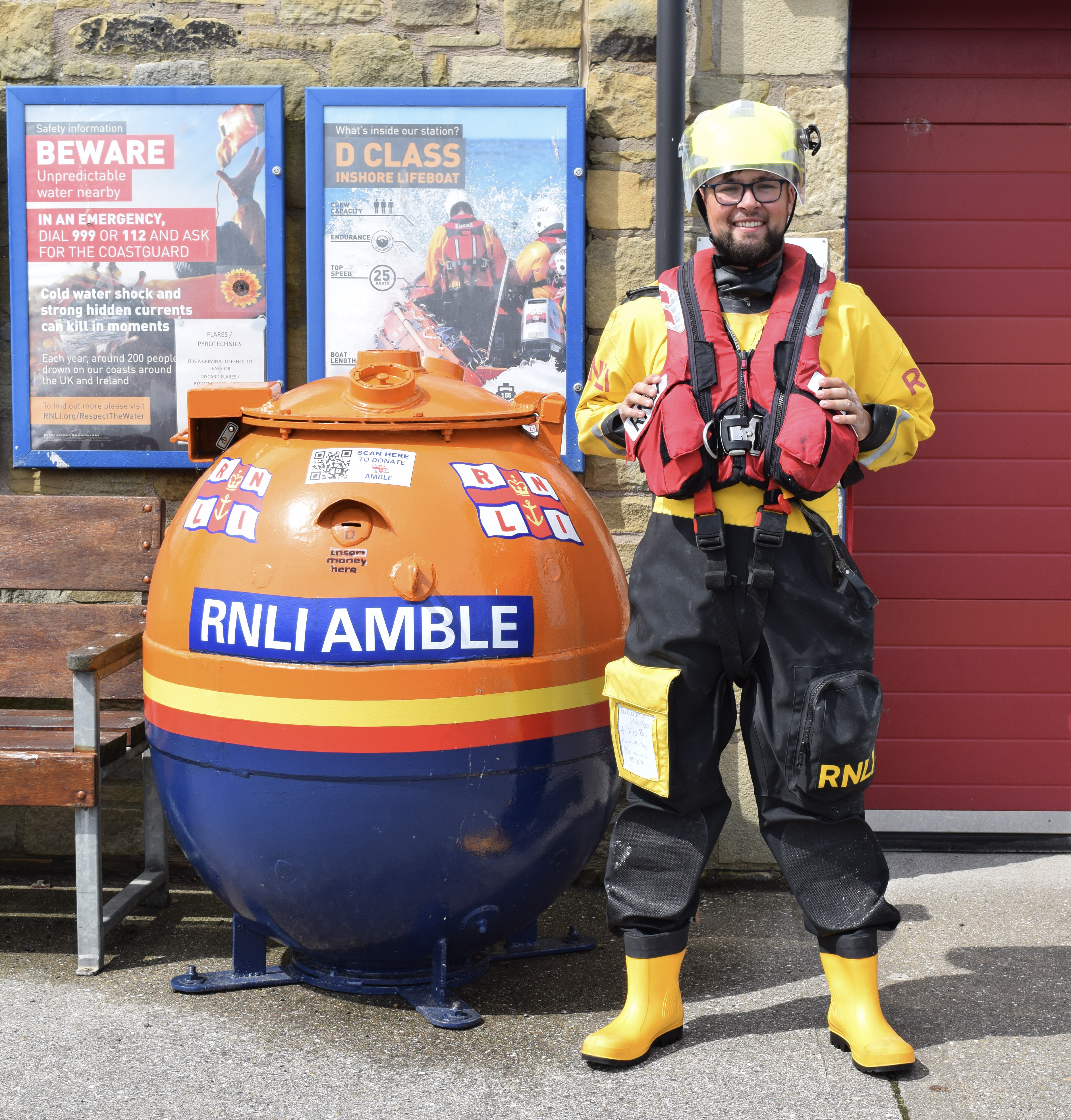 This image shows a young RNLI ILB lifeboat crew member standing in full gear outside of Amble Lifeboat Station. To the left of the crew member, a redundant wartime mine that has been repurposed and painted with RNLI paint and logos, stands nearby. The weather is bright and sunny.