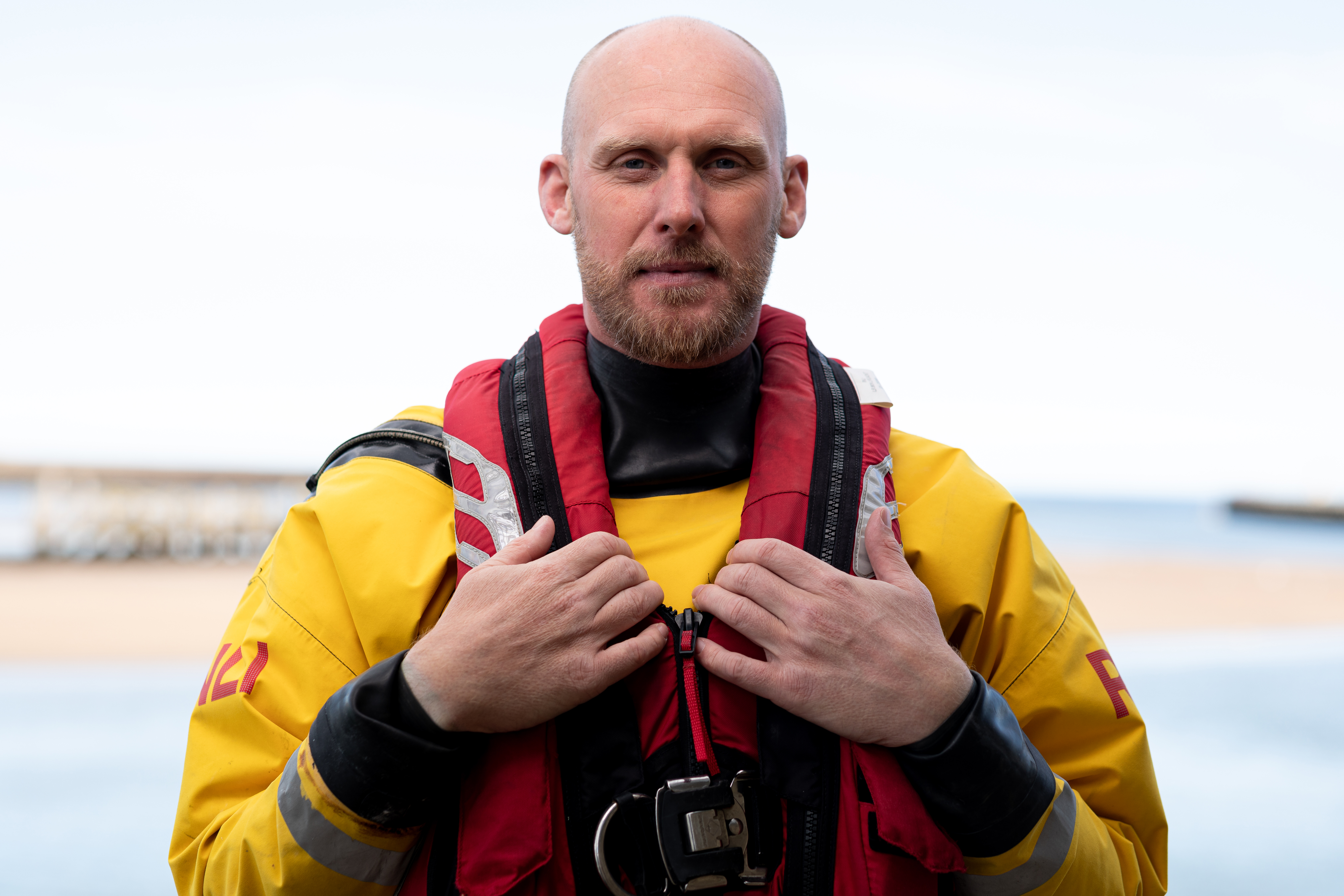 This image shows Amble's RNLI in-shore lifeboat helm Aaron Armstrong standing in full lifeboat gear. Aaron's hands rest on his floatation device, located around his neck and torso. The weather is cloudy but dry.