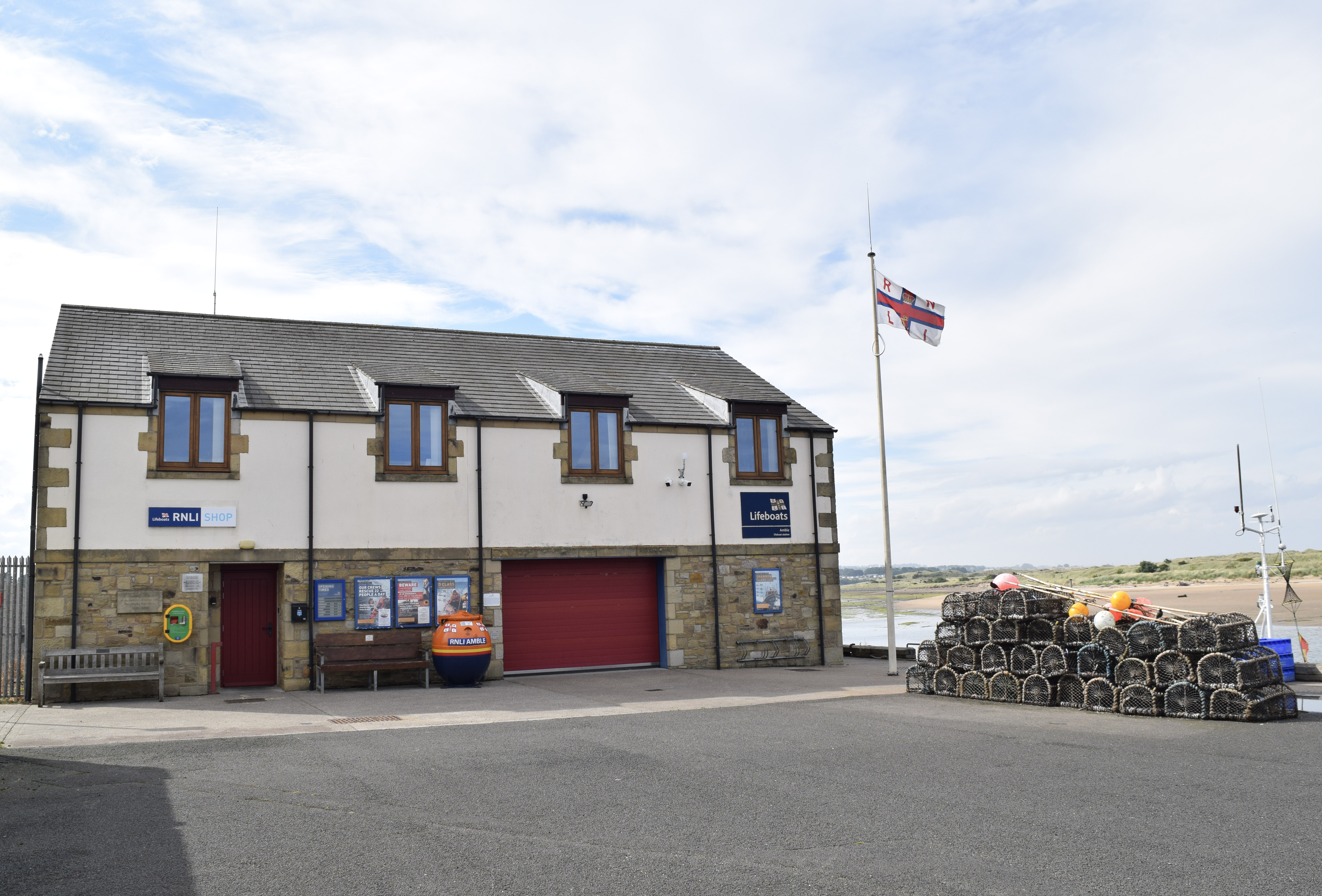 This image shows the RNLI Amble lifeboat station with a RNLI flag fluttering in the breeze nearby. Some of the nearby harbour and the opposite edge of the estuary can be seen in the background. The weather is bright and sunny.