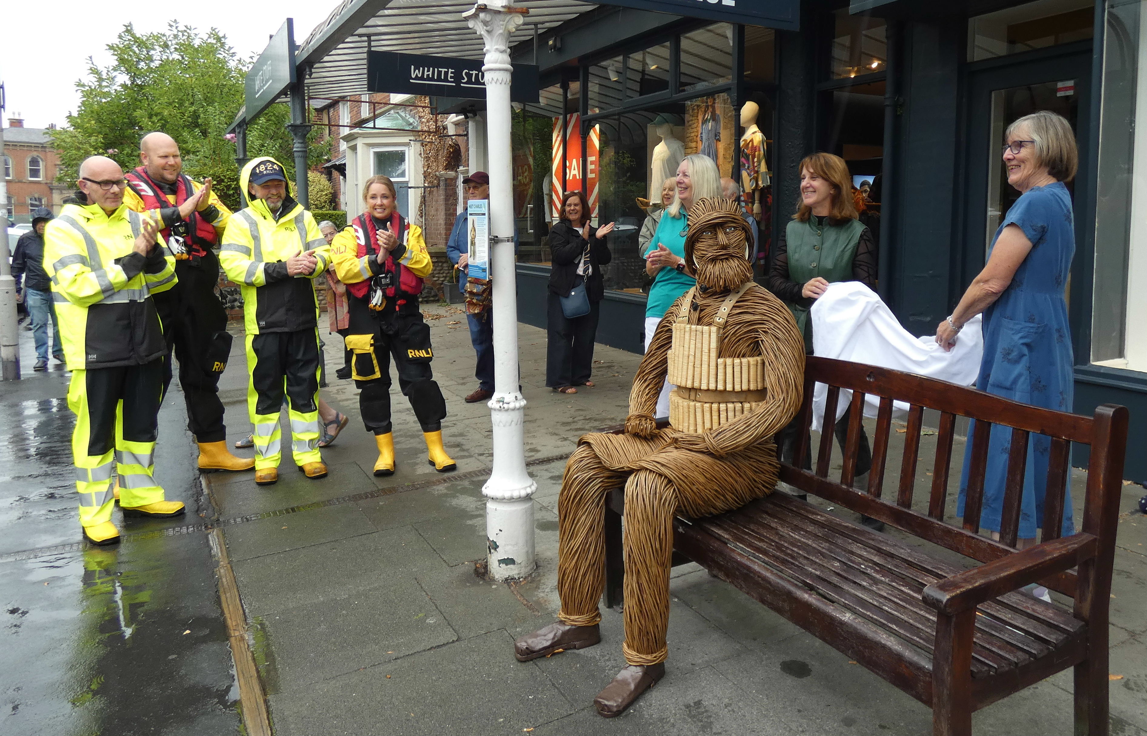 two ladies holding a cover watched by a Lifeboat Crew with the wicker man on the bench