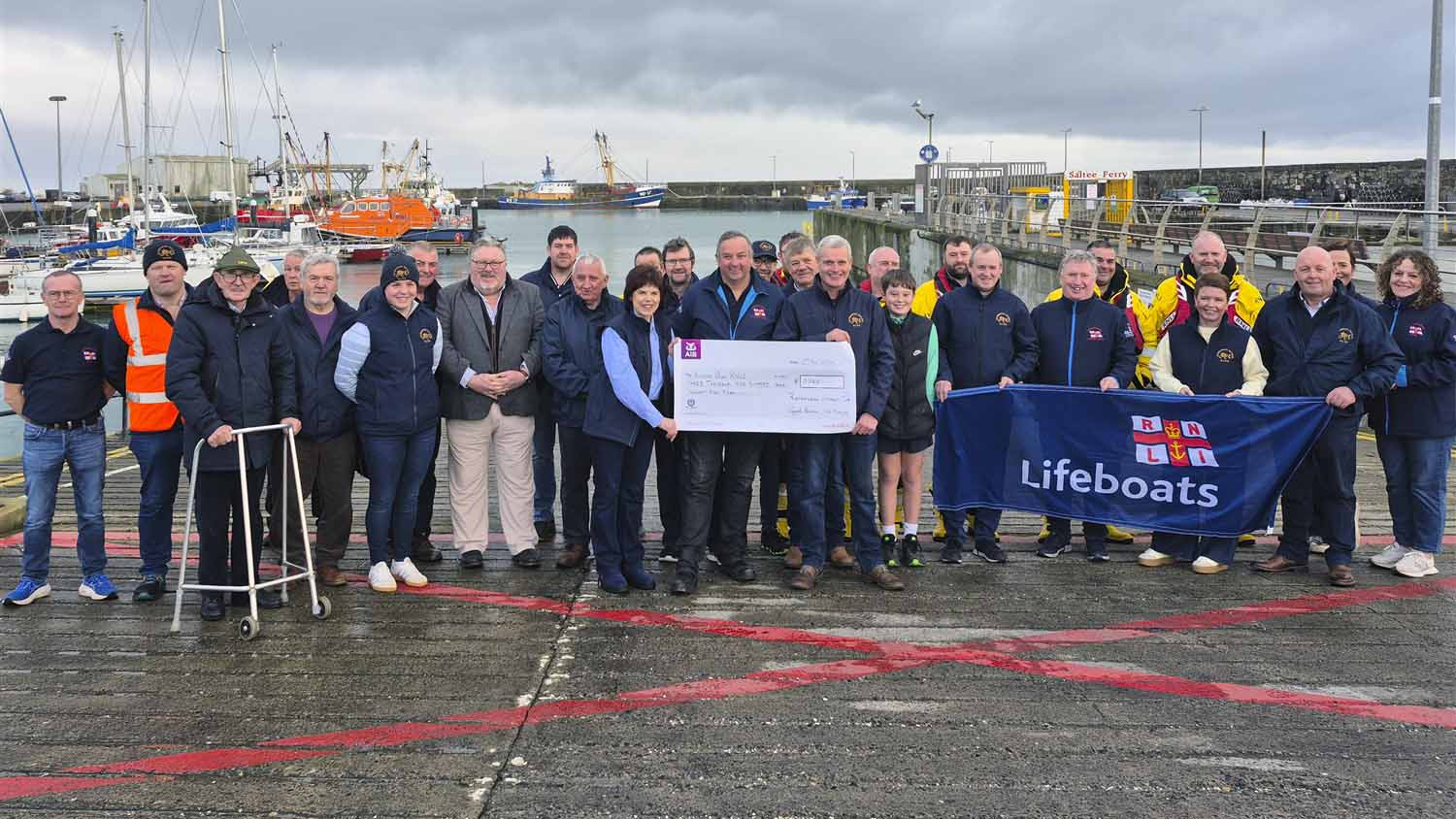 Group photo of Rathangan Vintage Club and crew members standing on the slip in Kilmore Quay with the lifeboat moored in the background