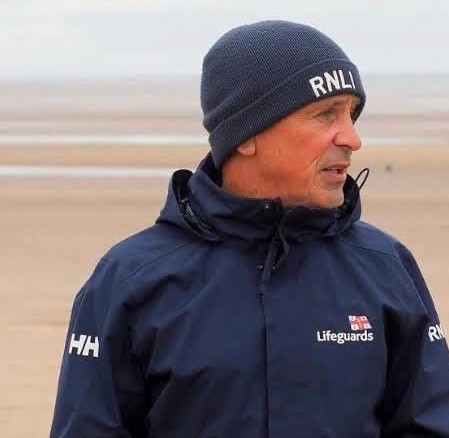 Richard on the beach on overcast day wearing RNLI branded navy clothing and beanie hat.