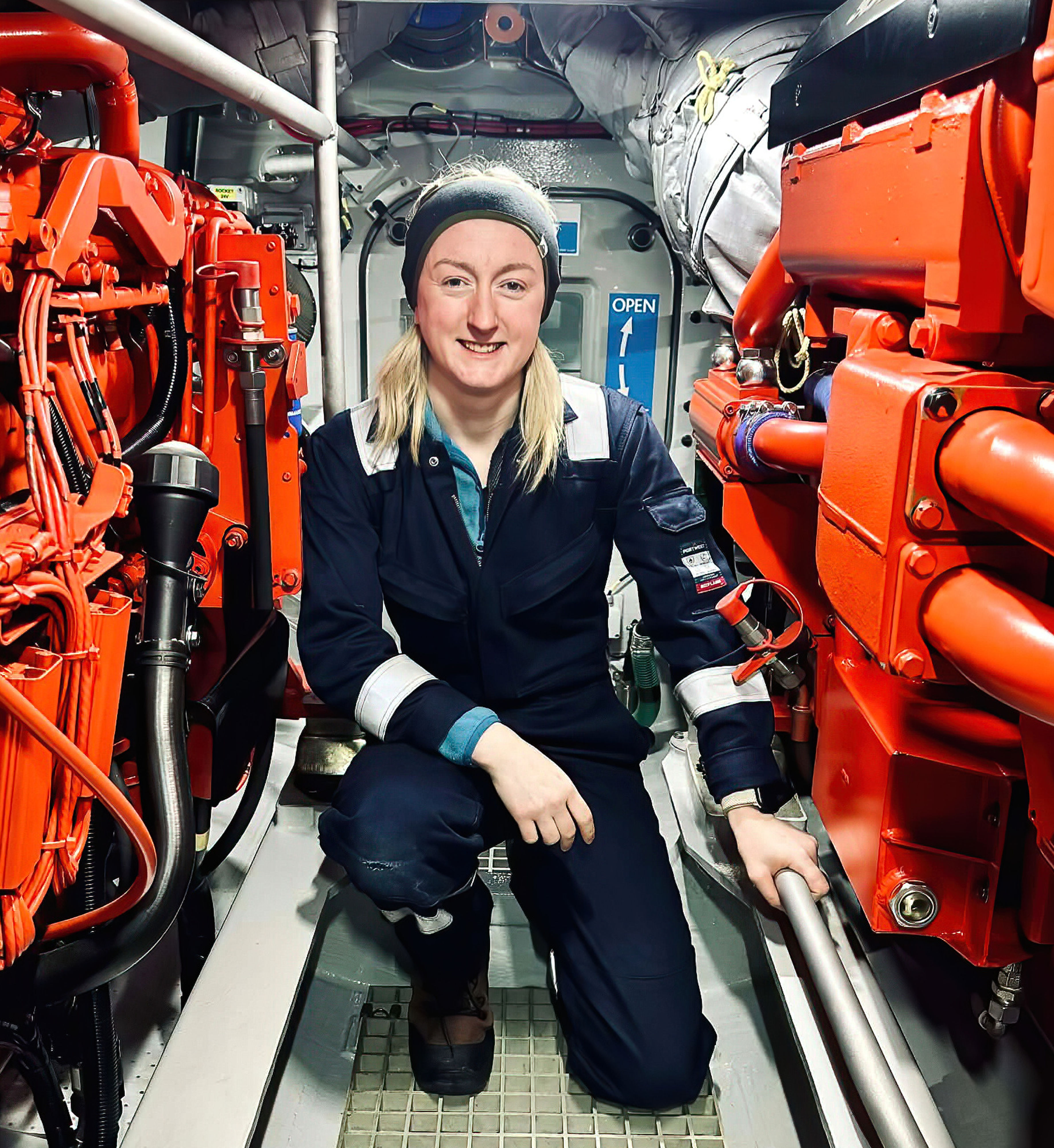 Lawrie kneeling in teh small space between the big orange engines in the cramped engine space below the waterline of the Shannon Class lifeboat