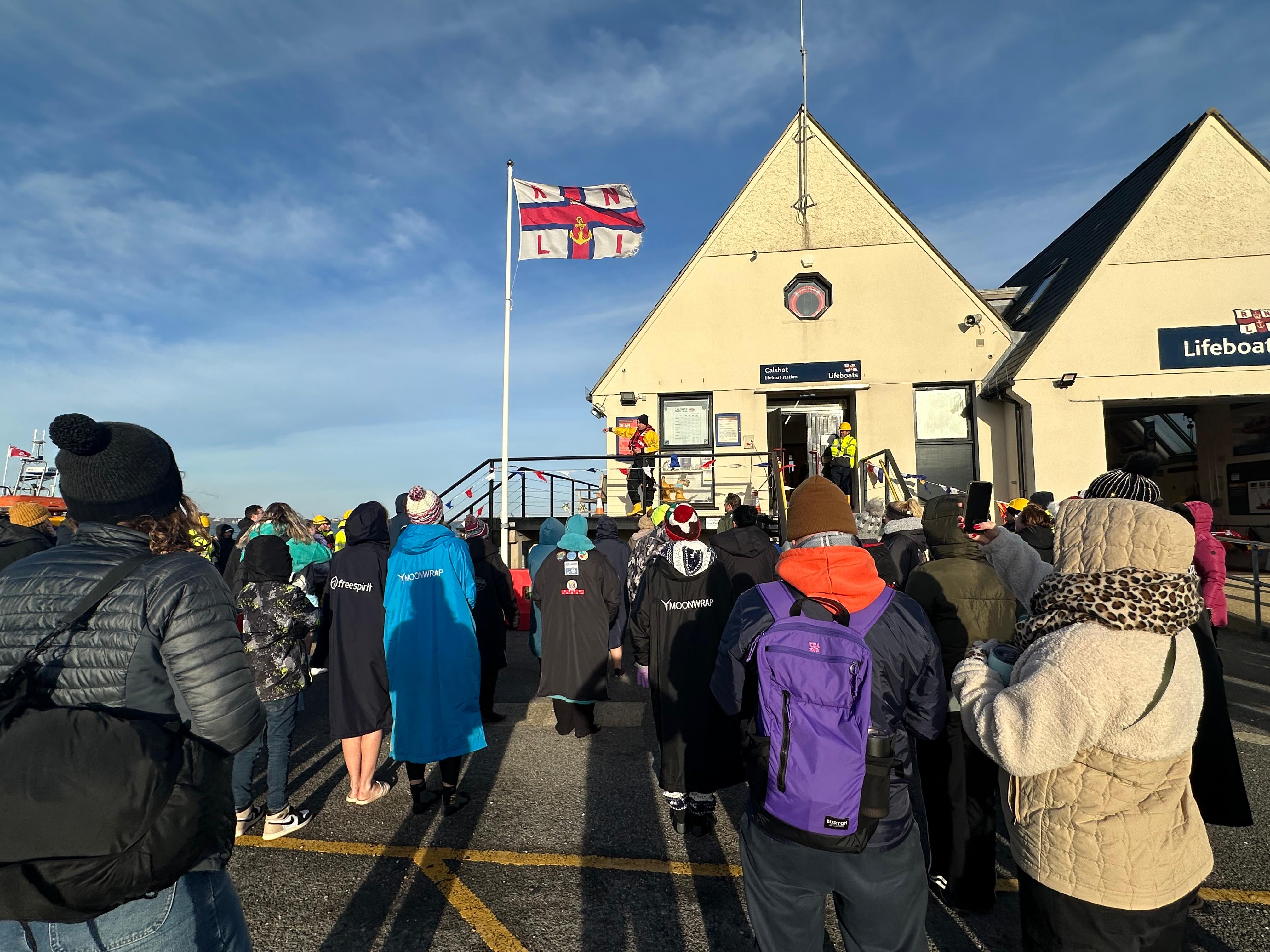 A man in an RNLI drysuit standing on a balcony speaking to a group of people.