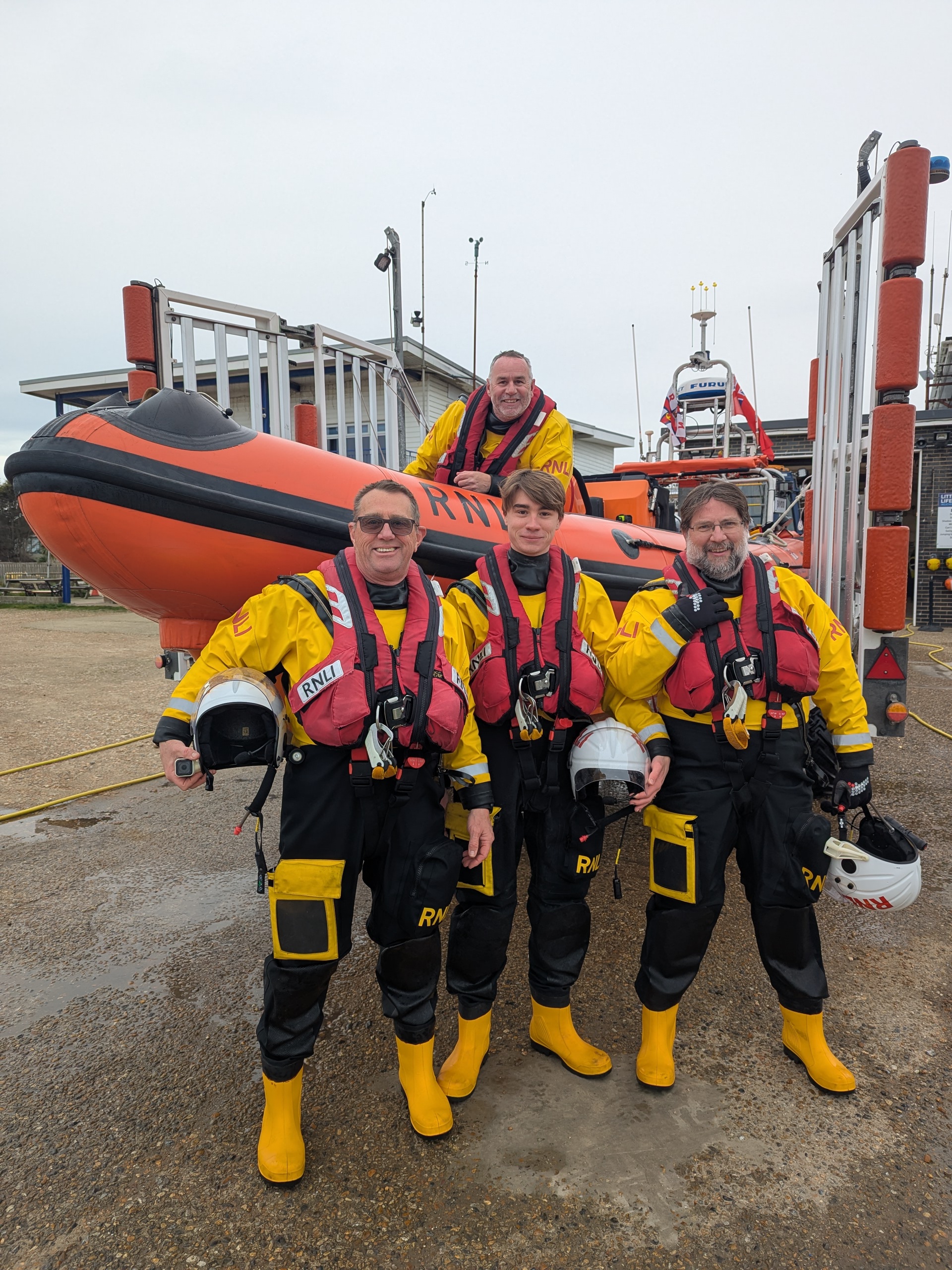 A smiling group of RNLI volunteer crew members in full yellow and black dry suits and red lifejackets in front of the current Littlestone lifeboat, the Jean McIvor