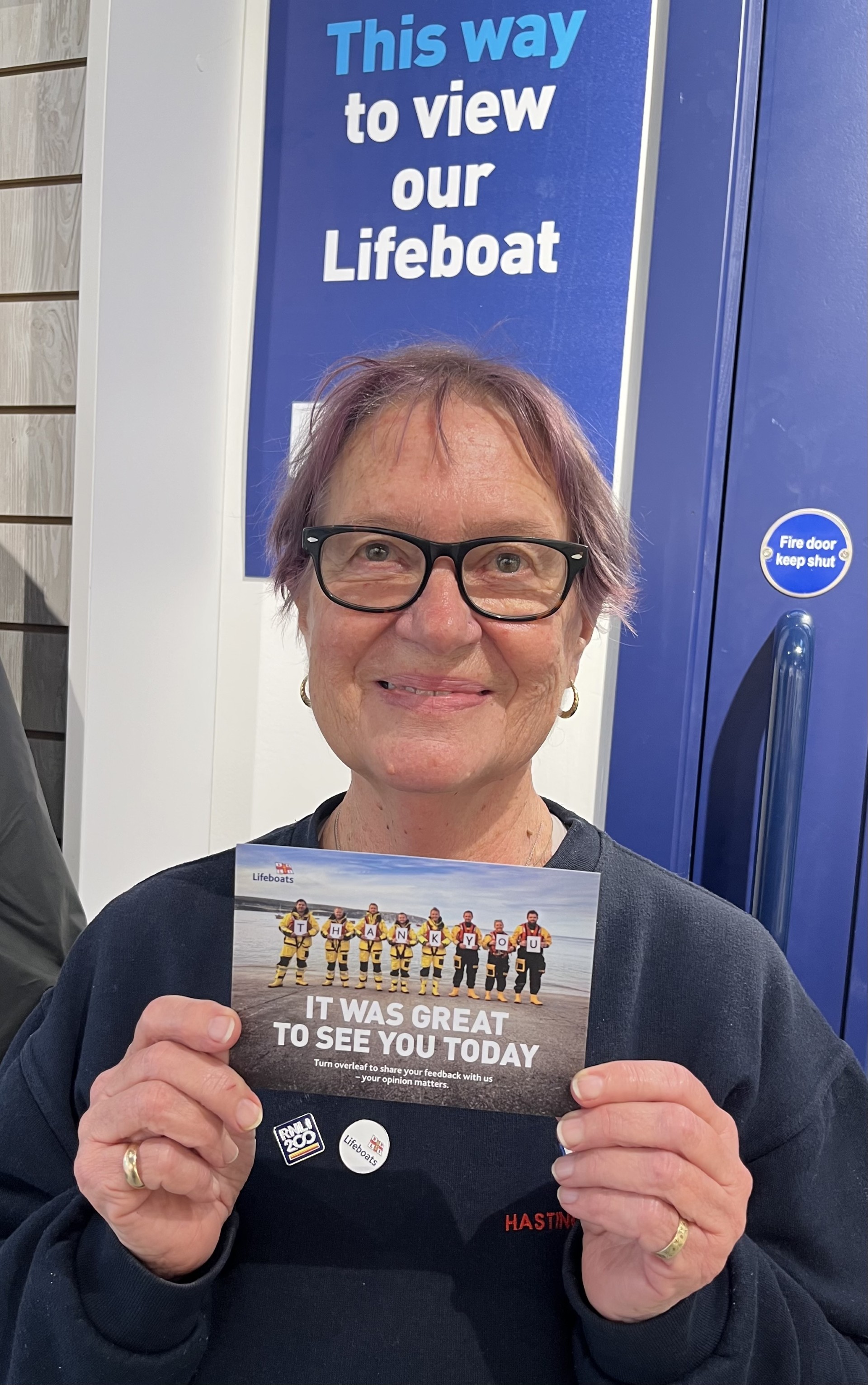 Hastings Lifeboat Station Visits Officer, Annette Veness, standing by the view the lifeboat sign, holding a postcard that states' it was great to see you today' featuring a lifeboat crew holding individual signs making up the word THANK YOU