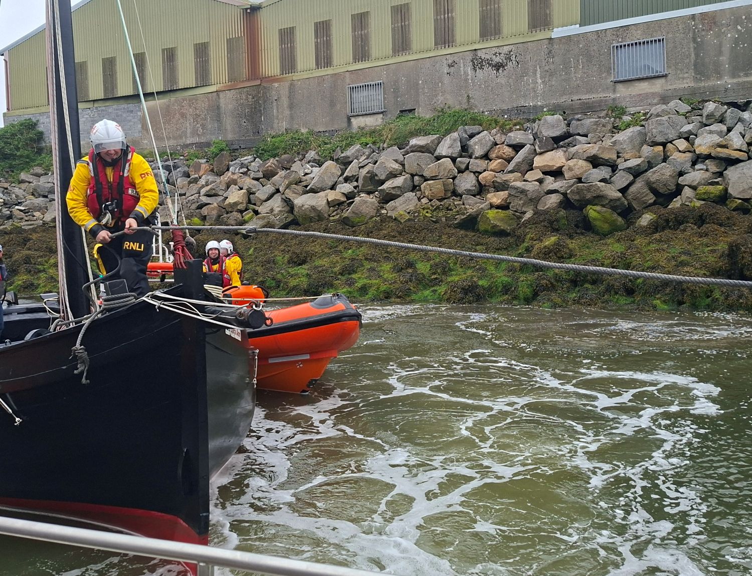 Crew member on the bow of a black sailing boat securing a rope as the boat is being tied up. Two lifeboat crew on an inshore lifeboat are just visible in the background on the right