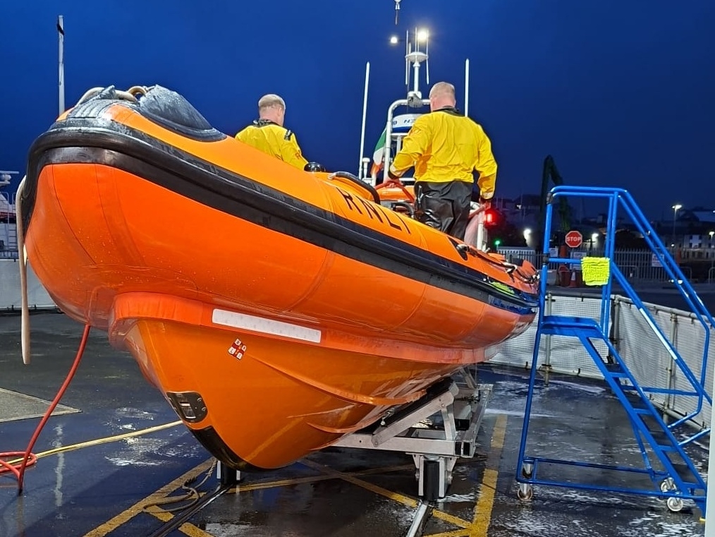 Two crew standing on the lifeboat which is on the cradle outside the lifeboat station. It is dark outside and the crew are facing away from the camera. There is water and suds on the ground around the lifeboat.