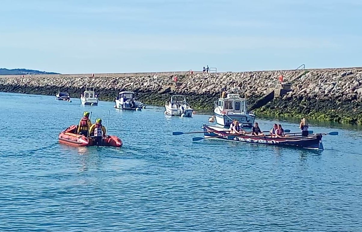 The wrapped gig boat rowing past Torbay RNLI's D class Inshore lifeboat in Brixham harbour