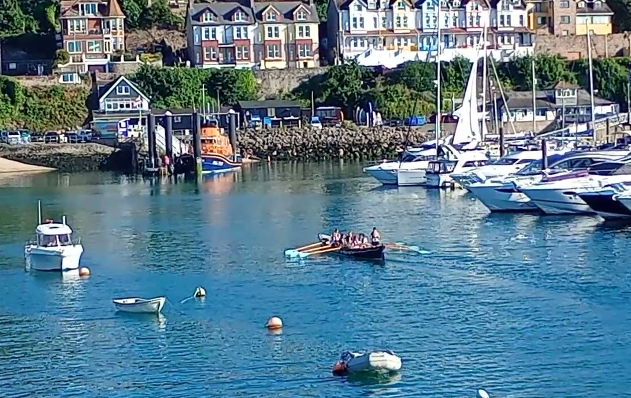 The wrapped gig boat approaching the slipway with Torbay Lifeboat Station in the background