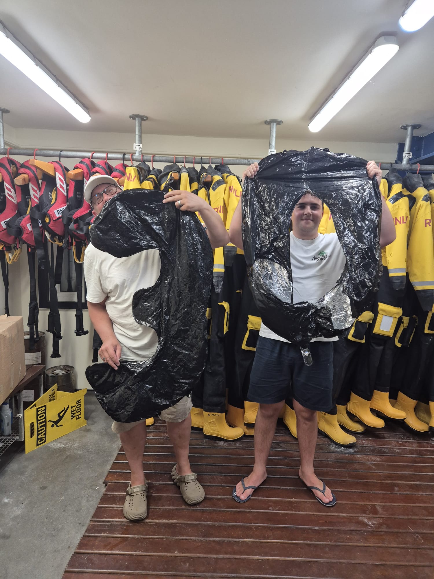 2 male volunteer crew members stand in the crew room at the lifeboat station, holding a number 3 and a number 0 balloon. They are surrounded by crew drysuits.