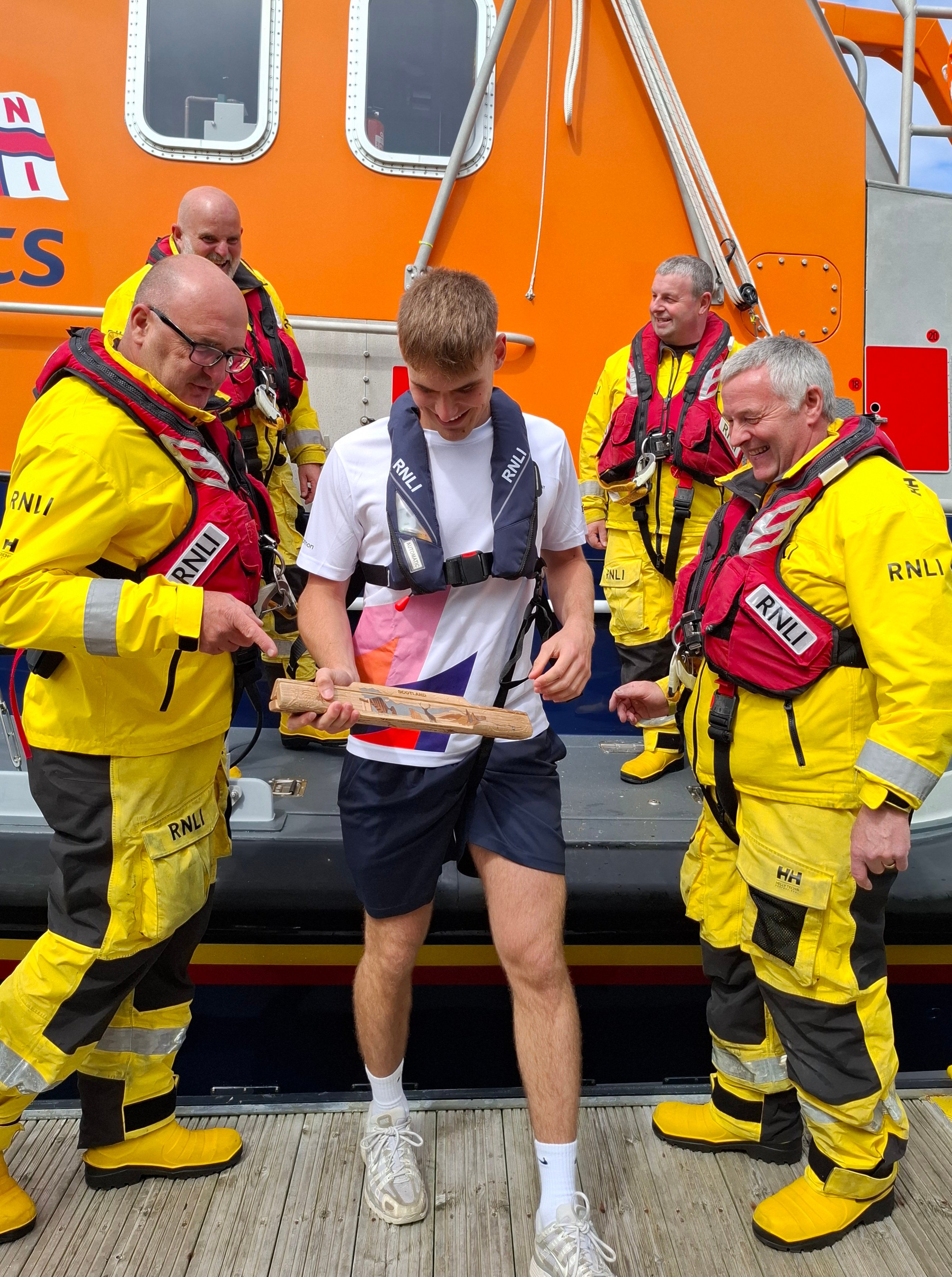 Four RNLI lifeboat crew, dressed in their yellow safety clothing, looking at the King's baton in the young athlete's hand before he takes it to the next carrier.