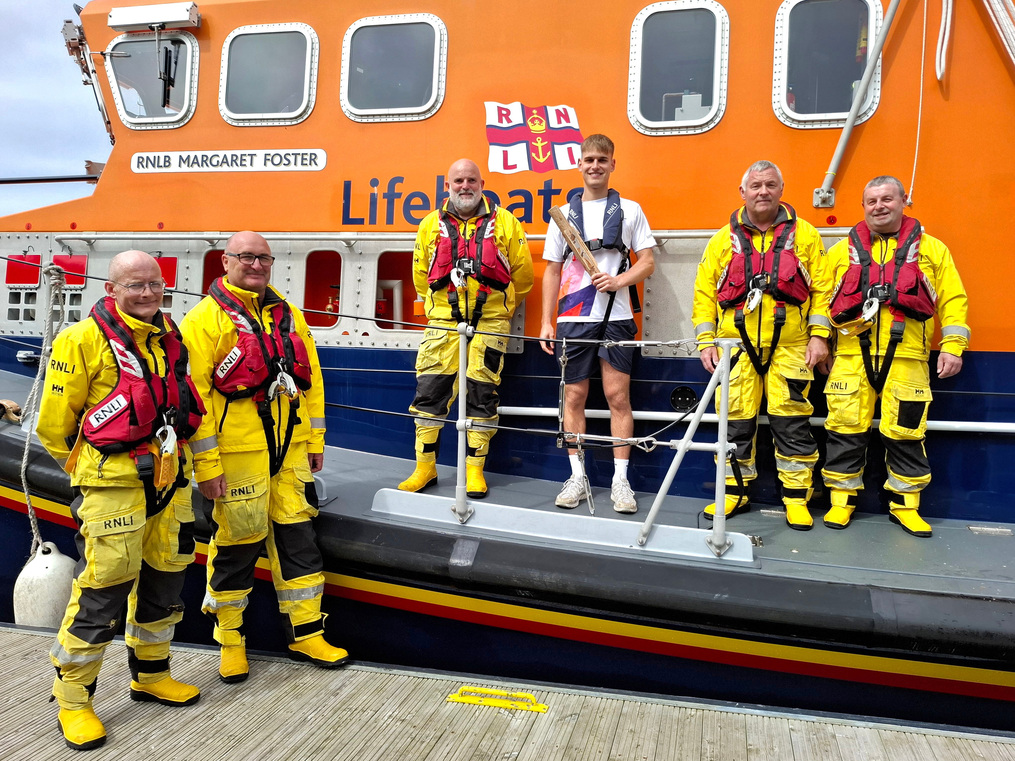 Five RNLI lifeboat crew, wearing their yellow safety clothing, by the lifeboat with young athlete holding the King's Baton.
