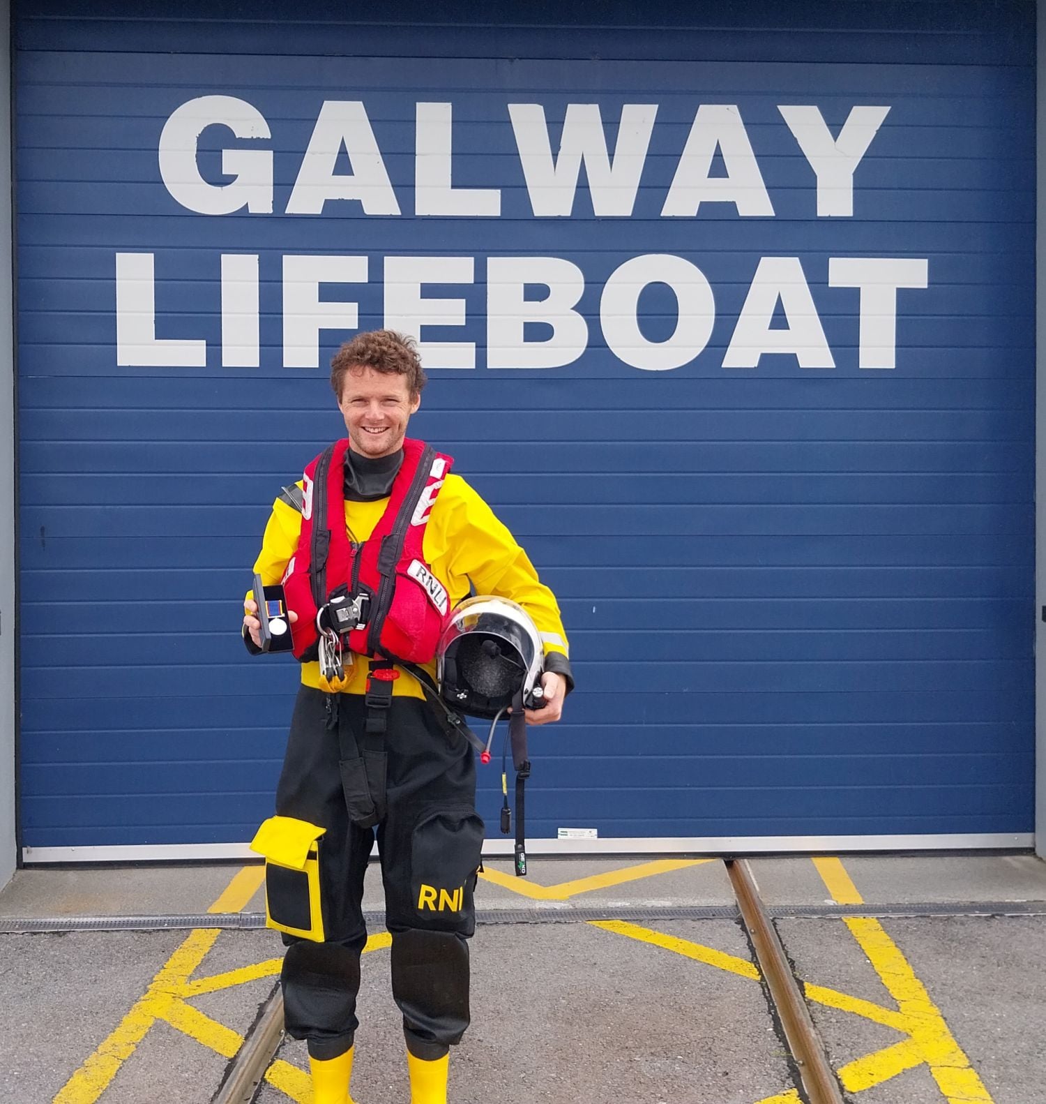 Crew in drysuit and lifejacket holding a 20 year medal in his right hand and his helmet in his left. Photo taken outside the shutter door of Galway Lifeboat Station