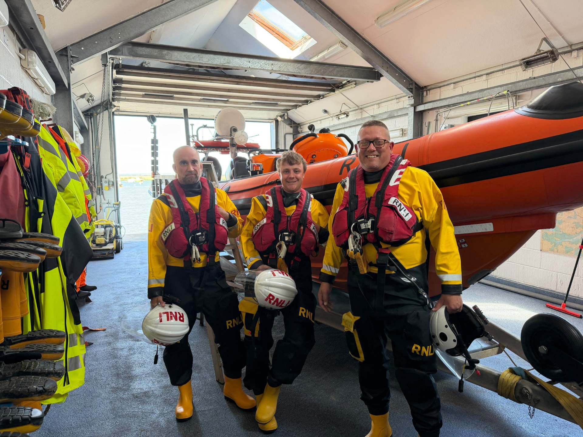 The photo is an image of 3 male lifeboat crew in drysuits and lifejackets, holding their helmets. They are stood in the boat house of the West Mersea lifeboat station, with the Atlantic 85 lifeboat named Just George behind them. They are smiling.