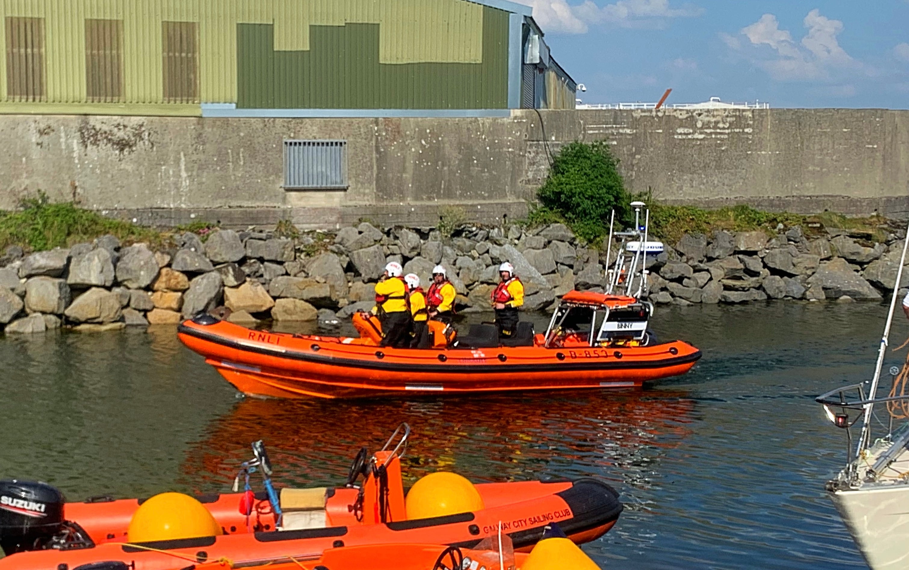 Atlantic 85 inshore lifeboat with 4 crew on board. The lifeboat is in the middle of a narrow channel with warehouses in the background and other boats tied up partially visible in the foreground.