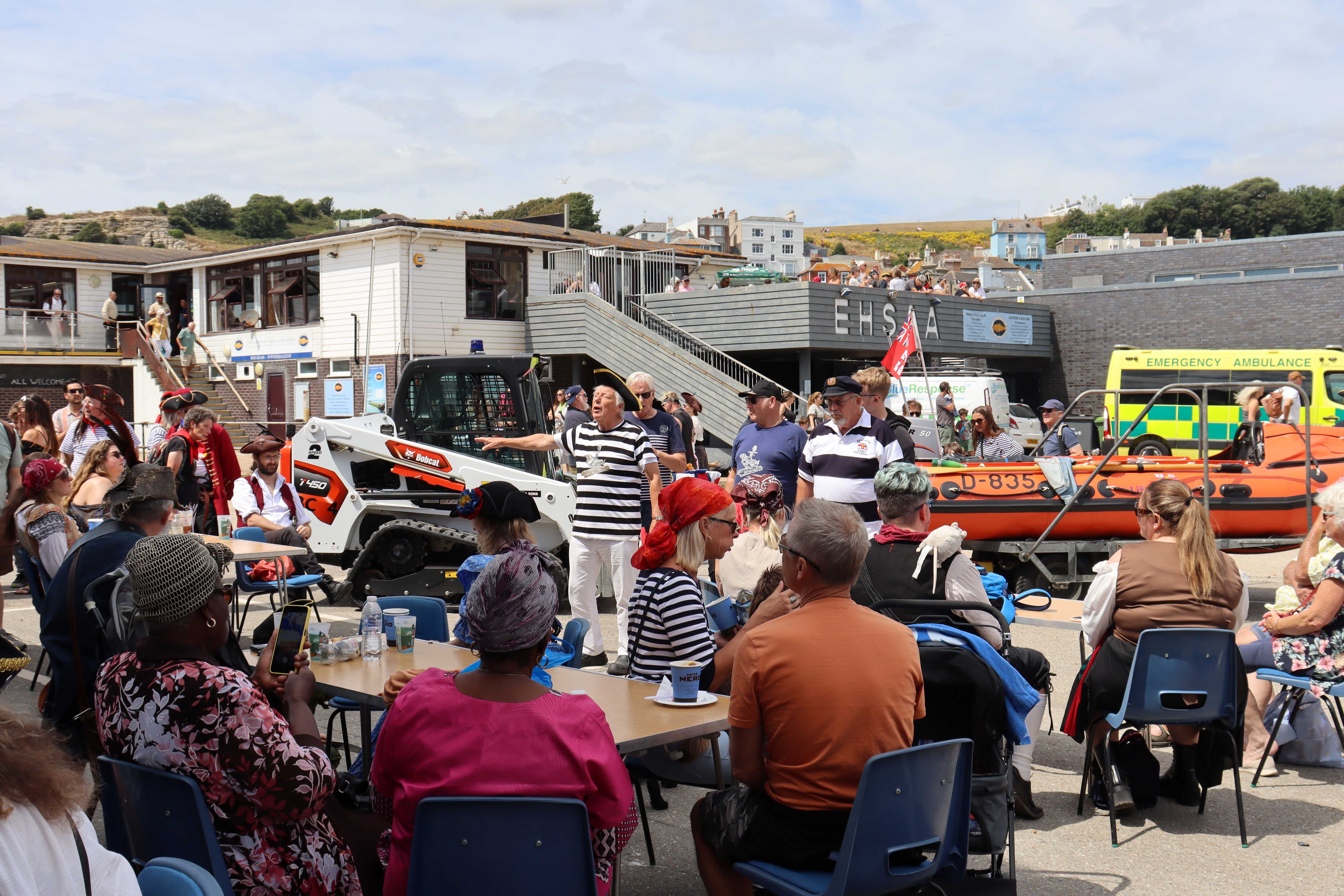 Groups of people sat at tables watching the shanty singers perform in front of an inshore lifeboat