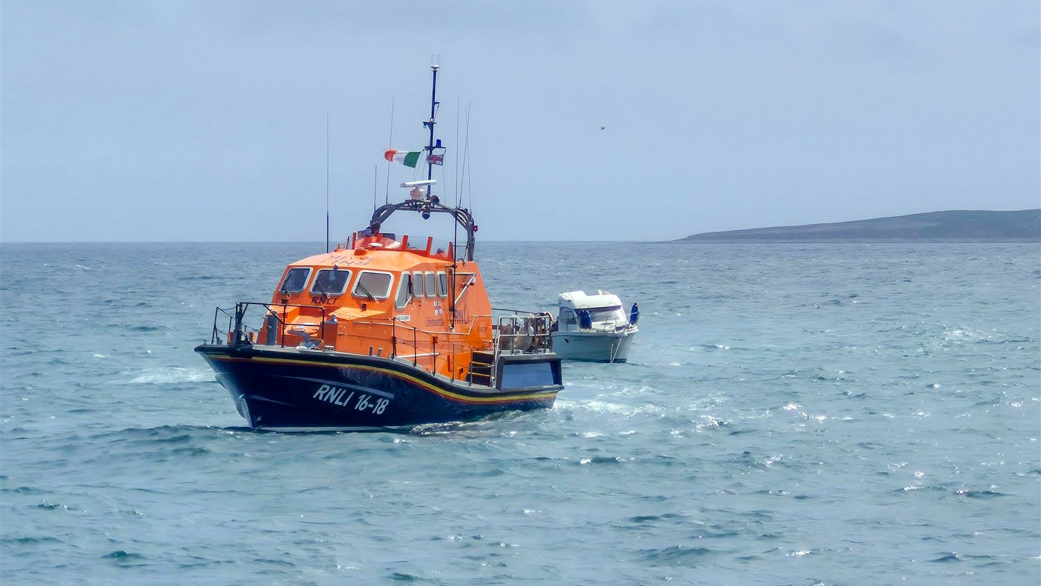 Slightly rippling sea and blue sky with Tamar class lifeboat in the foreground, a small white motorboat behind it at an angle connected by tow line, with the east end of Little Saltee island visible in the distant background