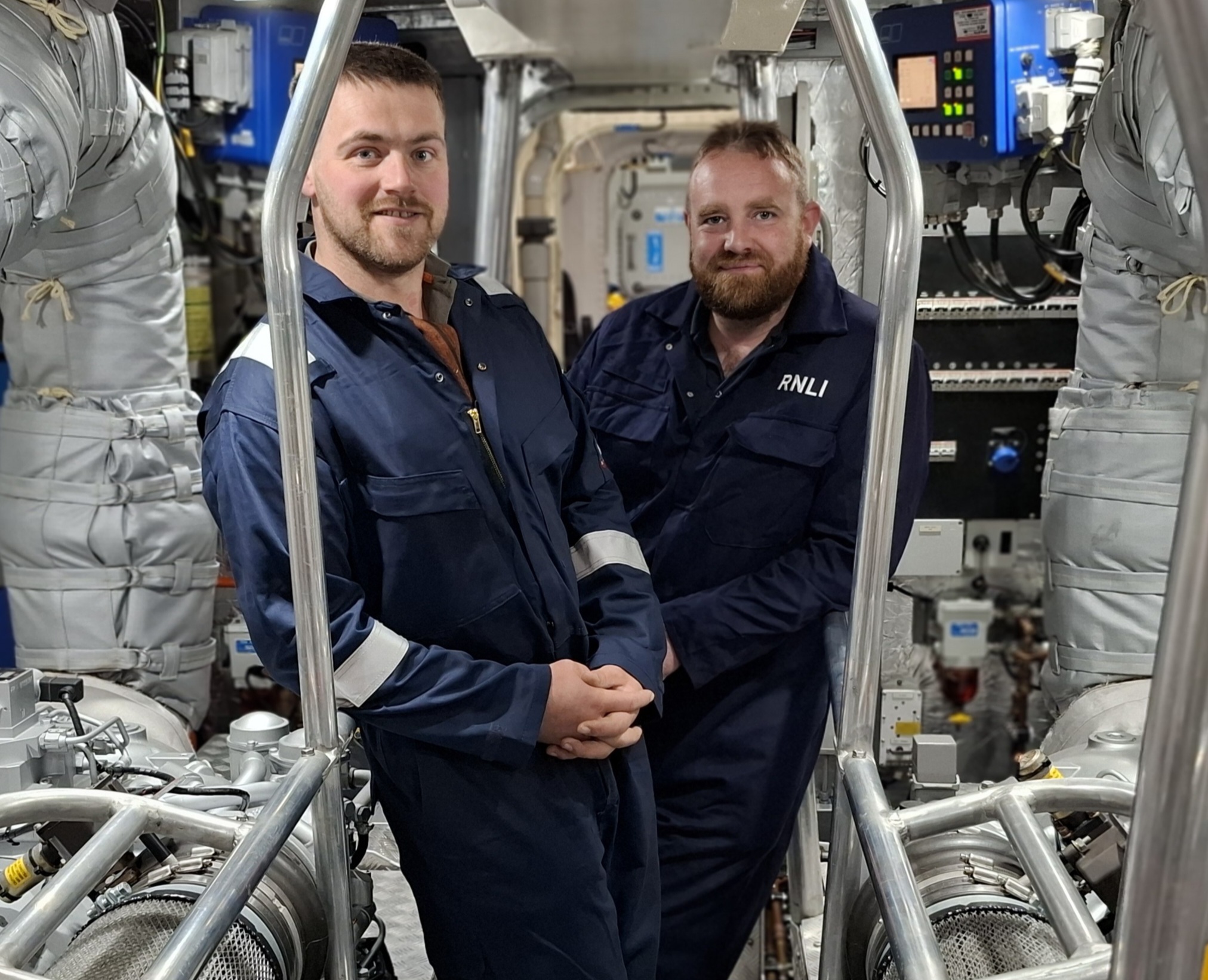 Two RNLI boat crew in navy overalls in the engine room of a lifeboat