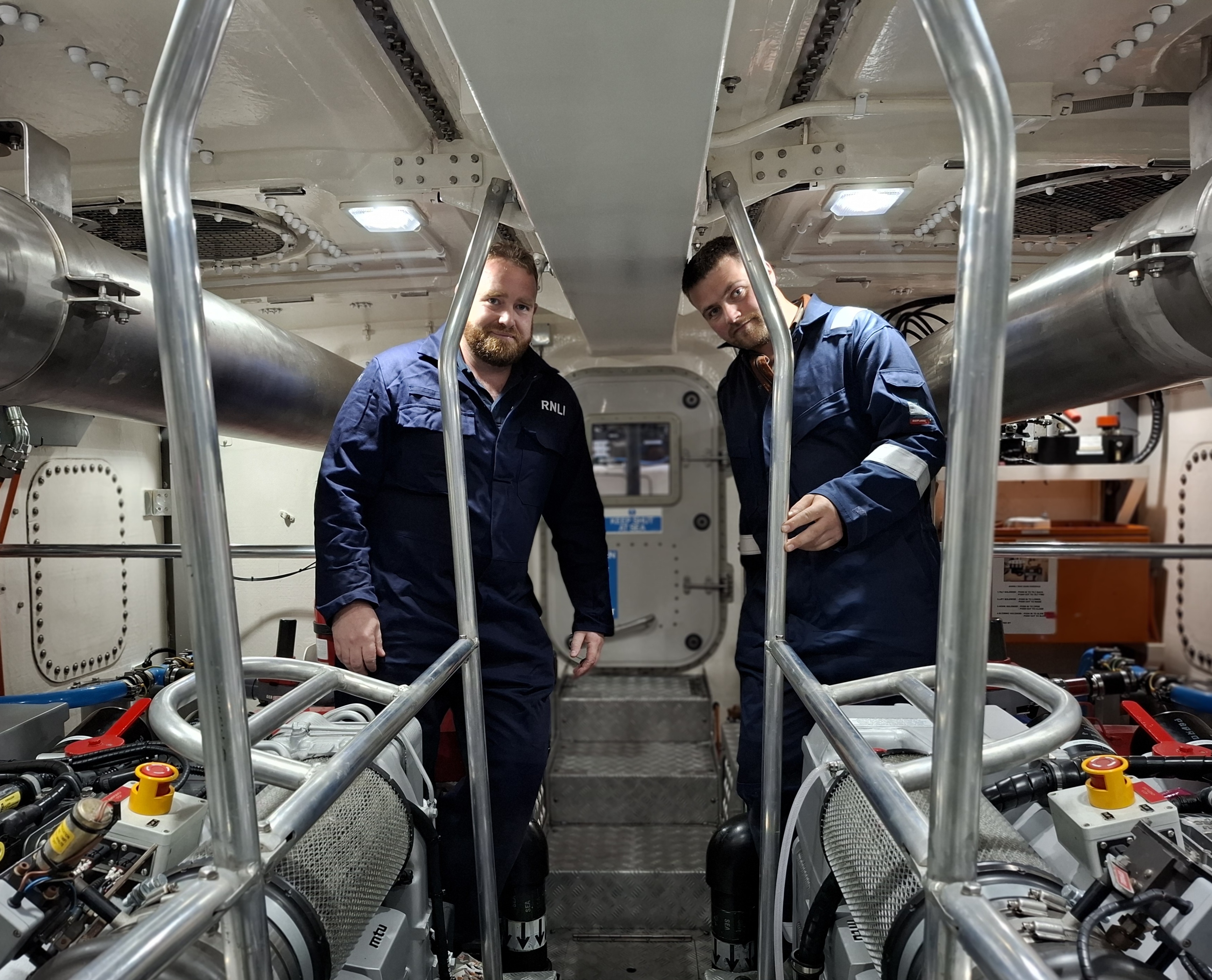 Two RNLI boat crew in navy overalls in the engine room of a lifeboat