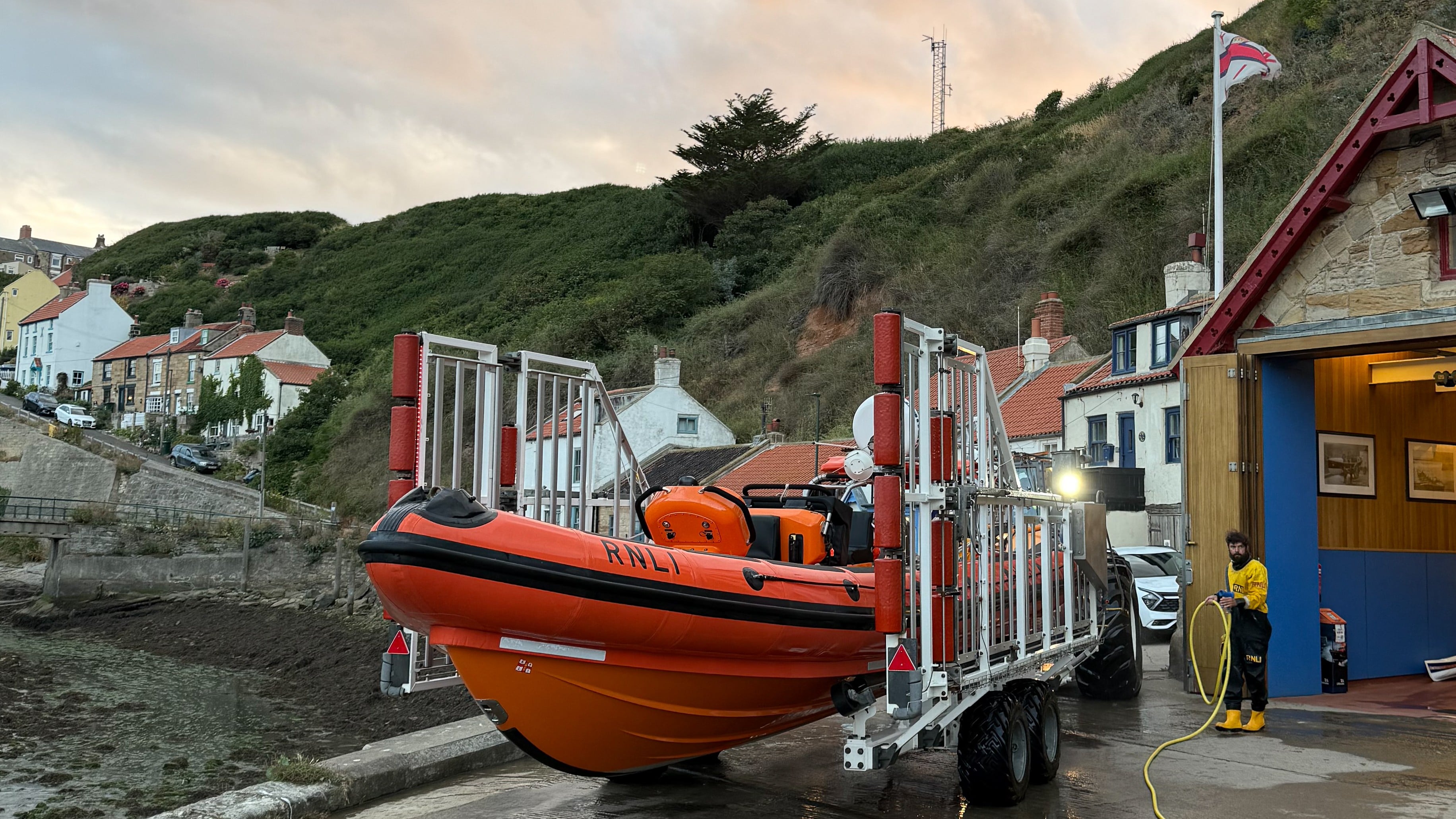 RNLI Atlantic 85 lifeboat B-897 Sheila and Dennis Tongue III sits on its recovery carriage outside the Staithes lifeboat station at dusk. The bright orange lifeboat is in the foreground, with the station building, harbour and steep green cliffs behind. An RNLI crew member is washing down equipment beside the boathouse as the lifeboat is made ready after training.