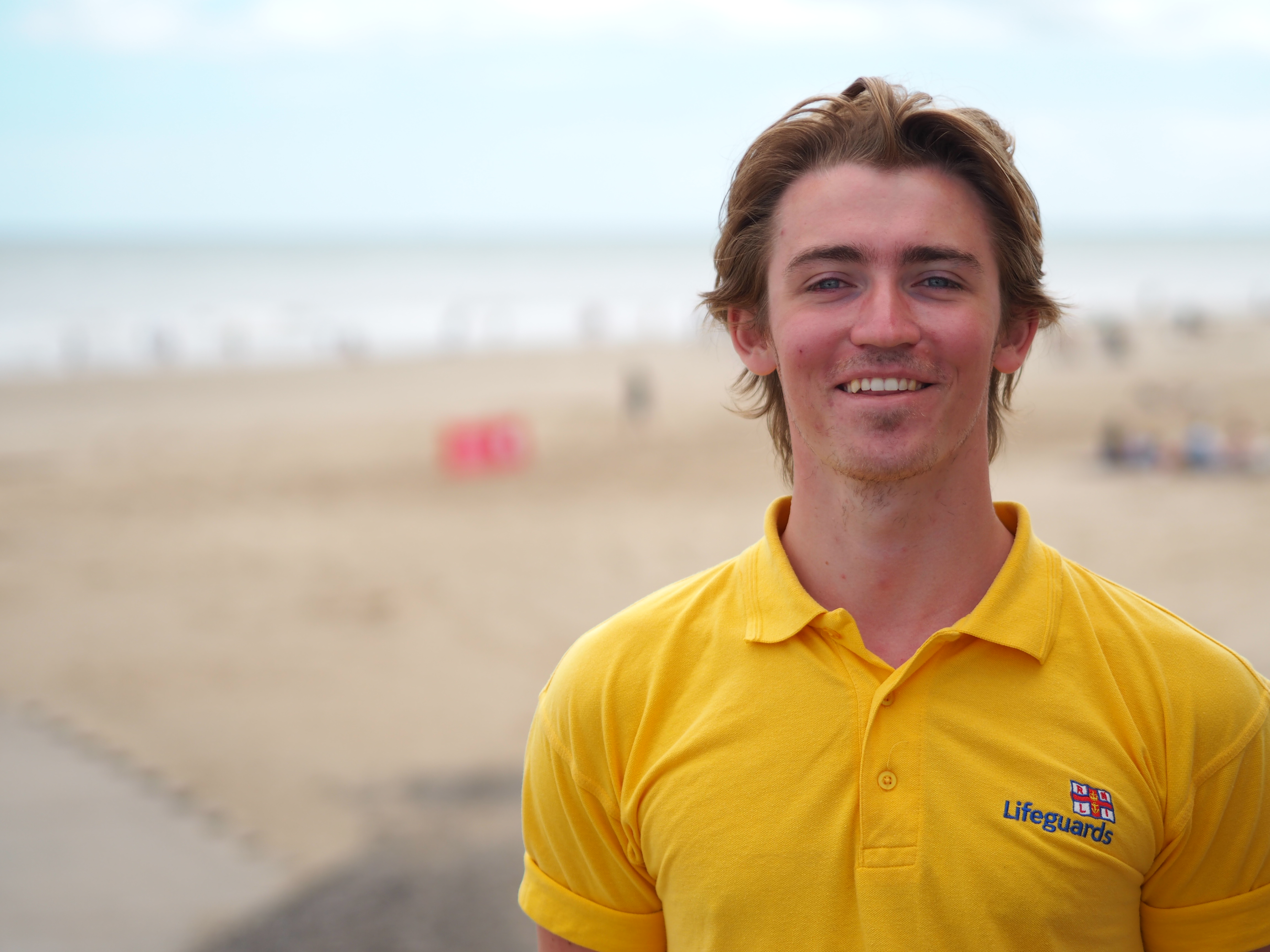 Liam Sumner, RNLI Senior Lifeguard, is dressed in a yellow t-shirt and is standing on a beach. He is serving in his eleventh season and has undertaken several notable rescues