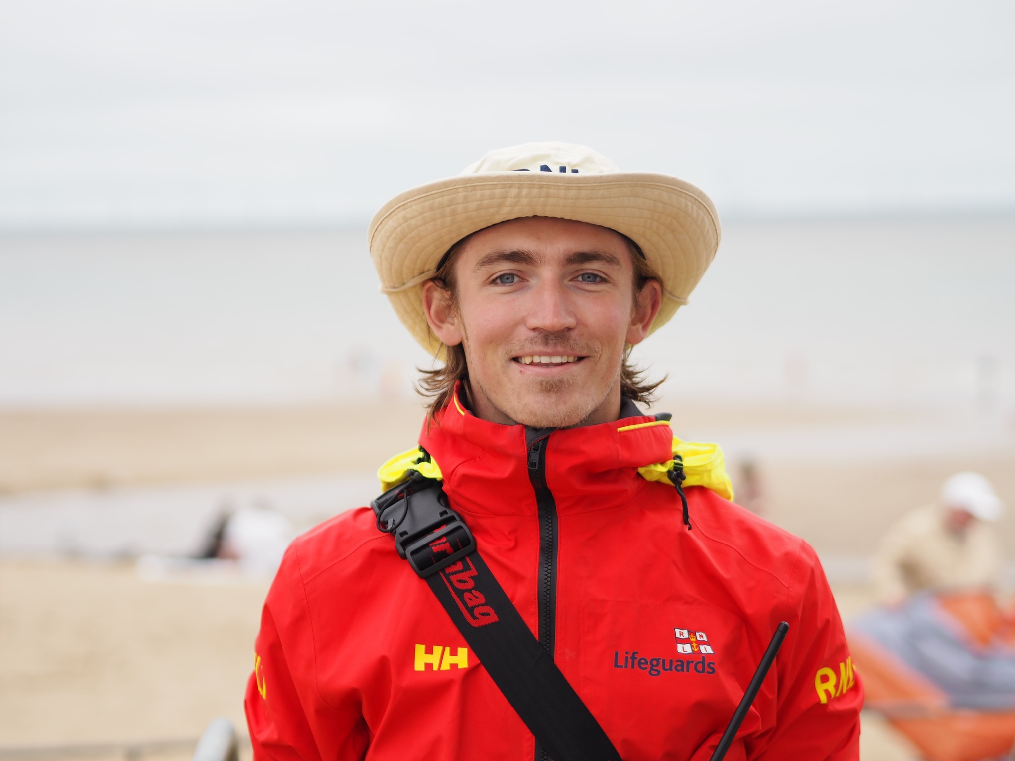 Liam Sumner, RNLI Senior Lifeguard, is dressed in an orange top and hat and is standing on a beach. He is serving in his eleventh season and has undertaken several notable rescues
